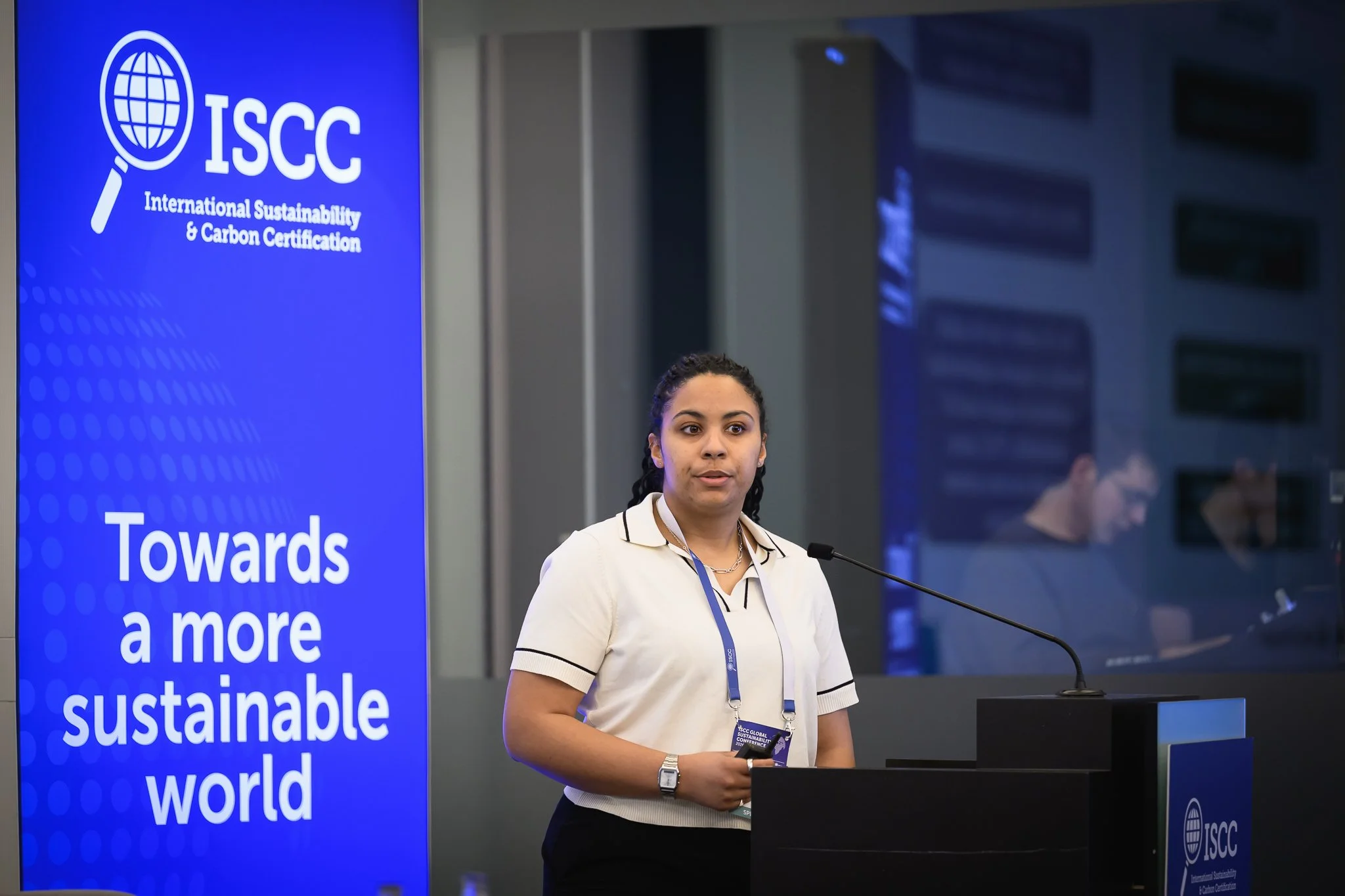 A woman in a light blouse stands at the lectern beside a blue ISCC banner.