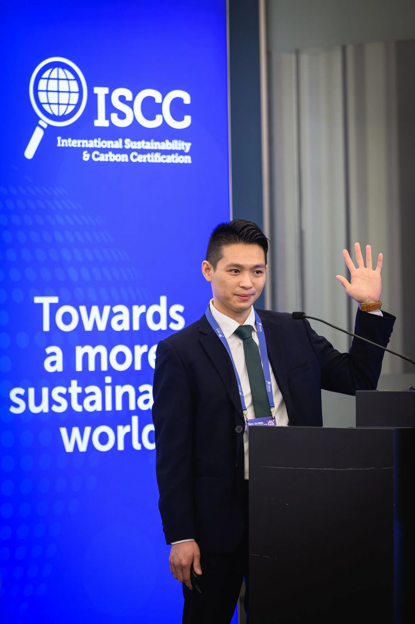 A man raises one hand while standing beside the lectern and an ISCC banner.