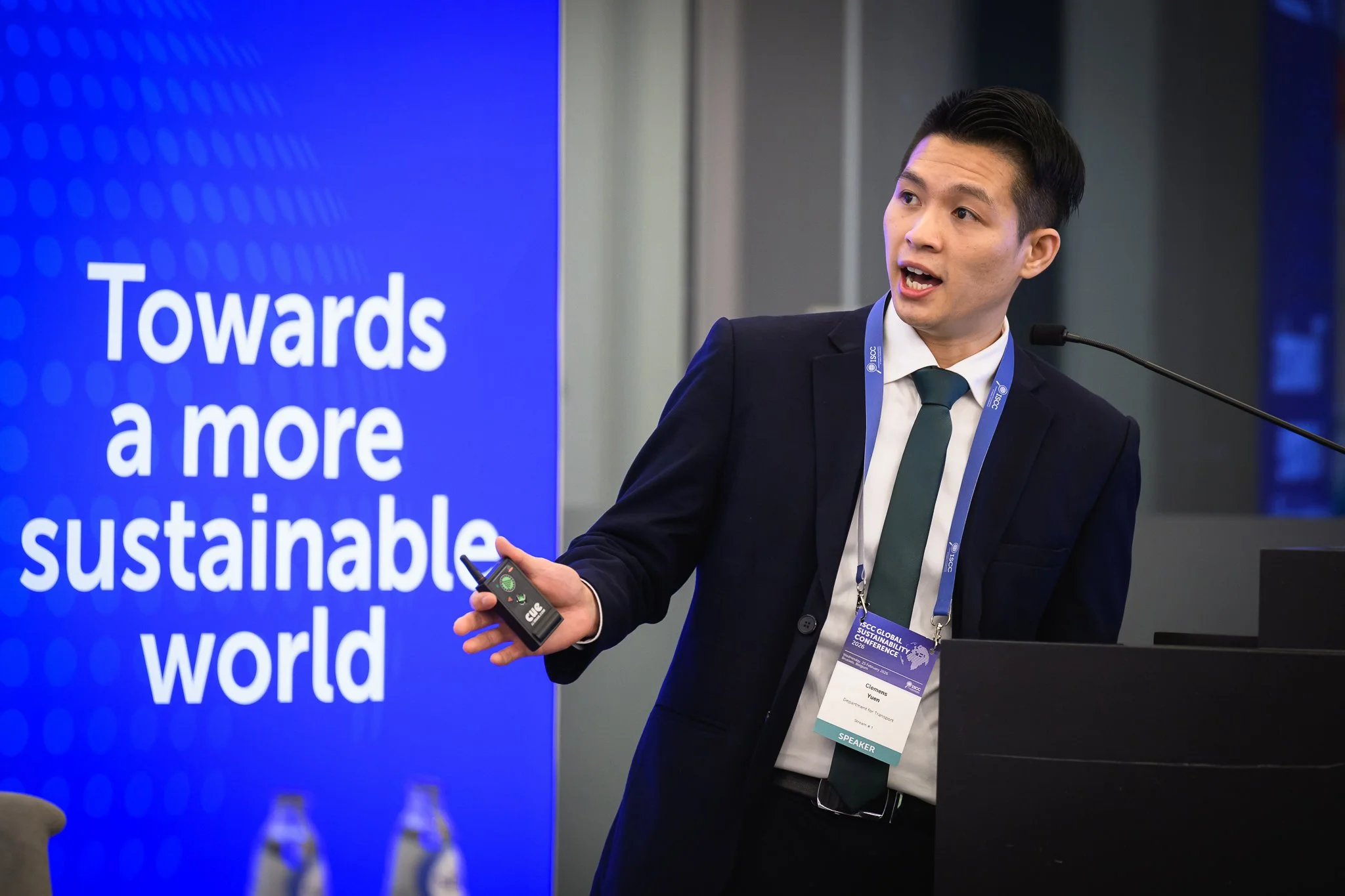 A man in a dark suit gestures with a remote control beside the conference lectern.