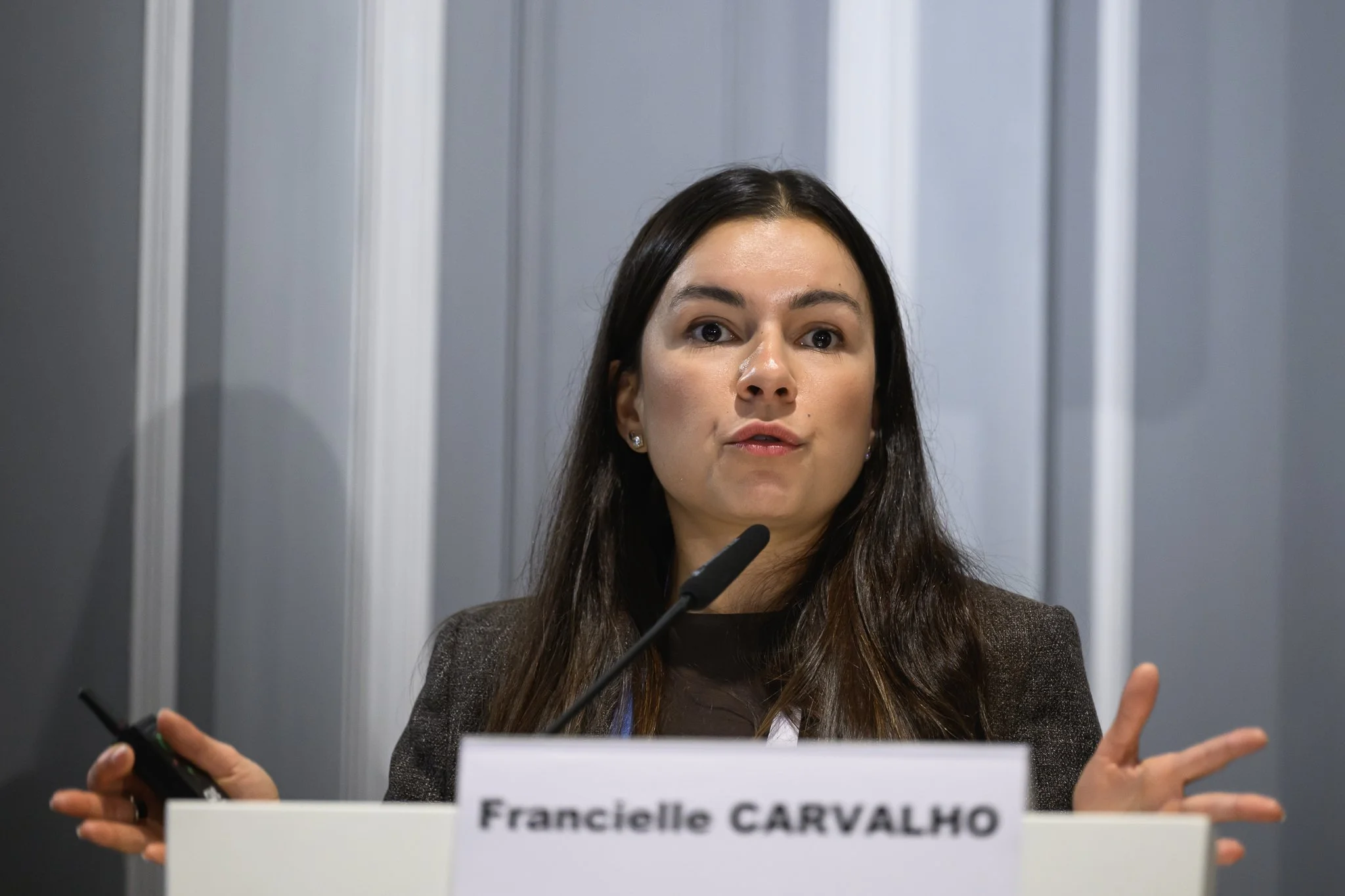 Francielle Carvalho speaks into the lectern microphone with both hands extended.