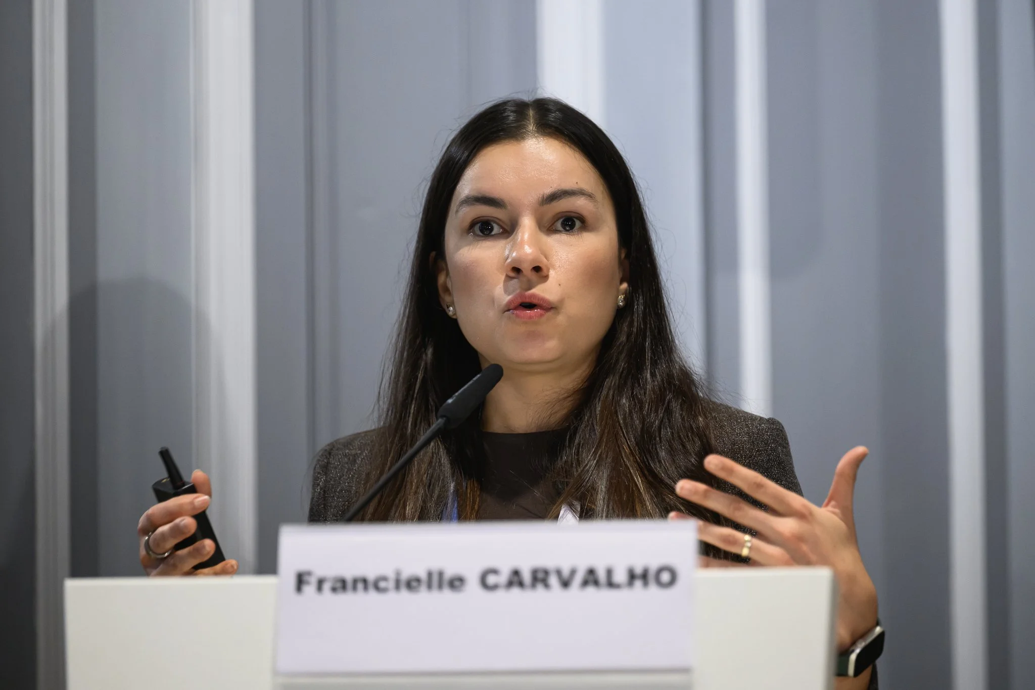 Francielle Carvalho gestures with a presentation remote while speaking at the lectern.