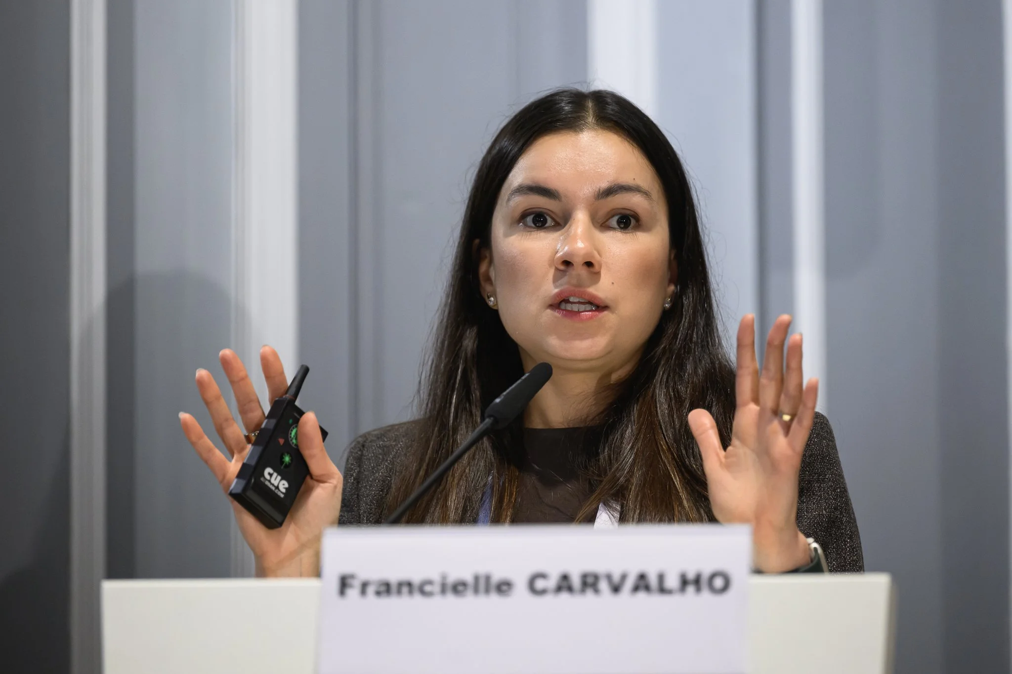 Francielle Carvalho raises both hands while speaking at a lectern bearing her name.