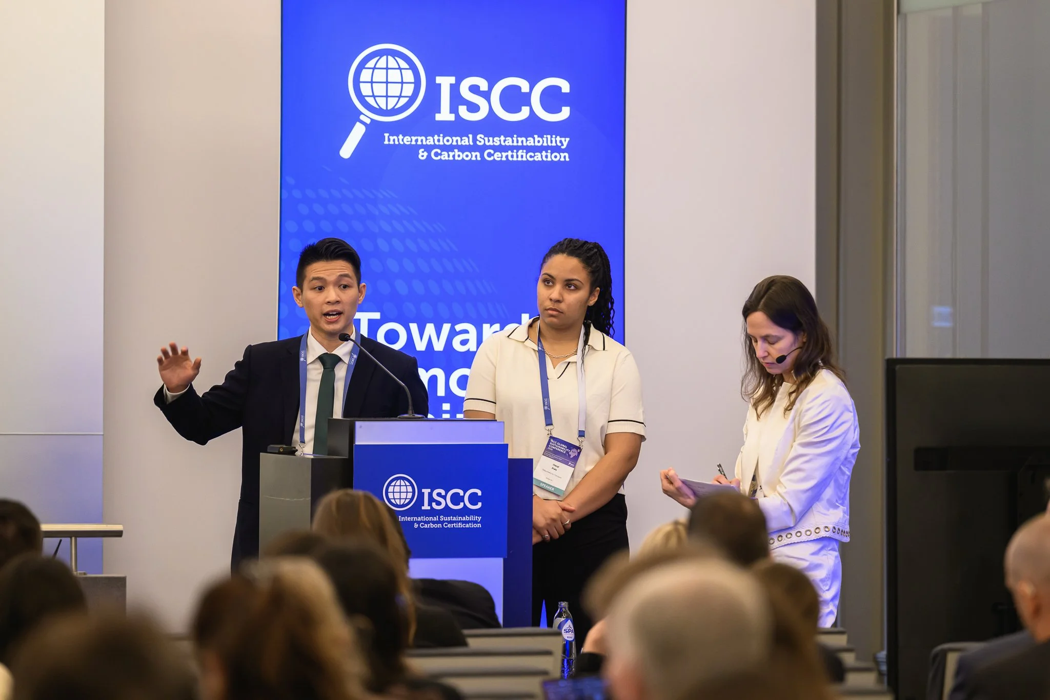 A man speaks at the lectern while two women stand beside him on stage.