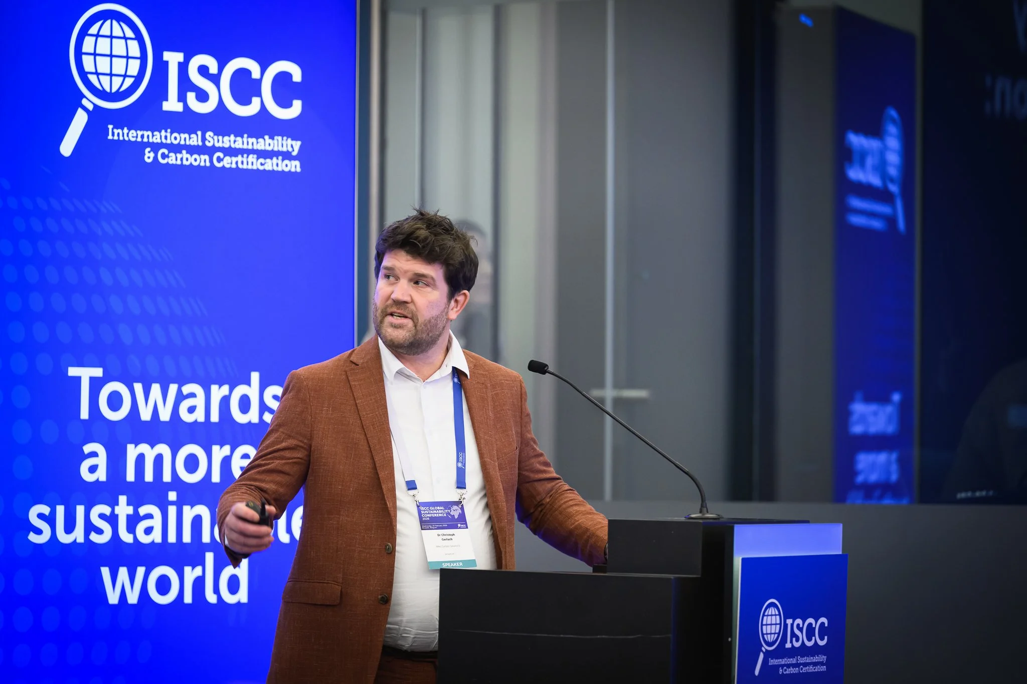 A bearded presenter in a brown jacket speaks beside an ISCC banner.