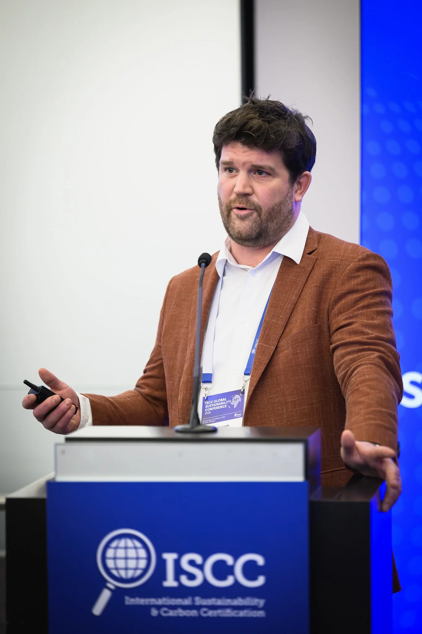 A man in a brown jacket gestures while presenting at the lectern.