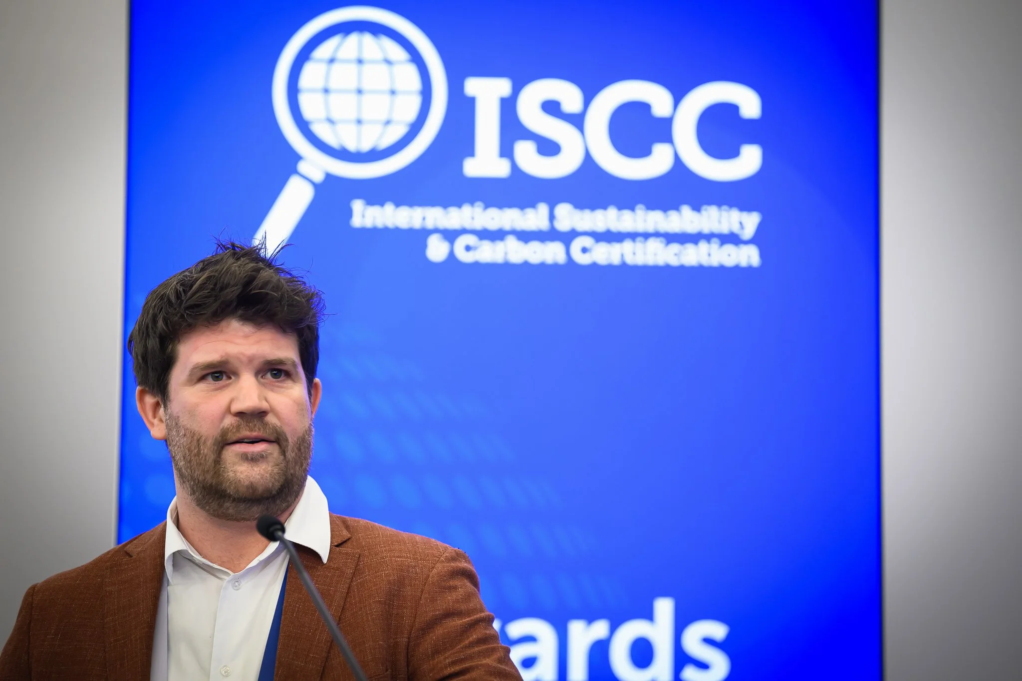 A bearded man in a brown jacket stands in front of a blue ISCC backdrop.