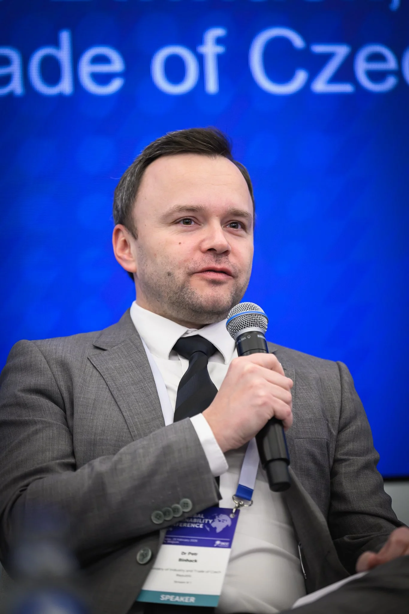 A seated man in a grey suit speaks into a handheld microphone.