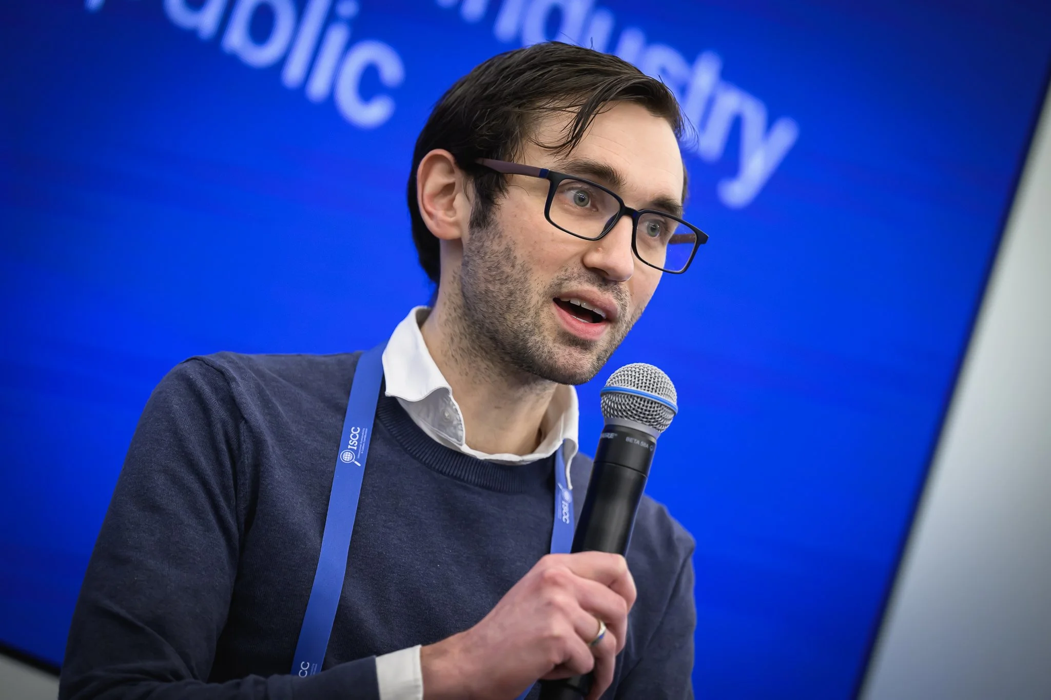 A seated man with glasses and a dark sweater speaks into a microphone.