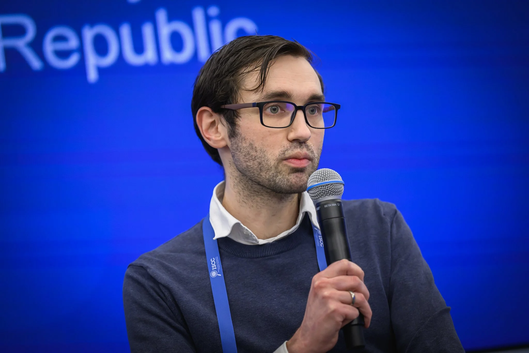 A man with glasses holds a microphone while seated on stage.