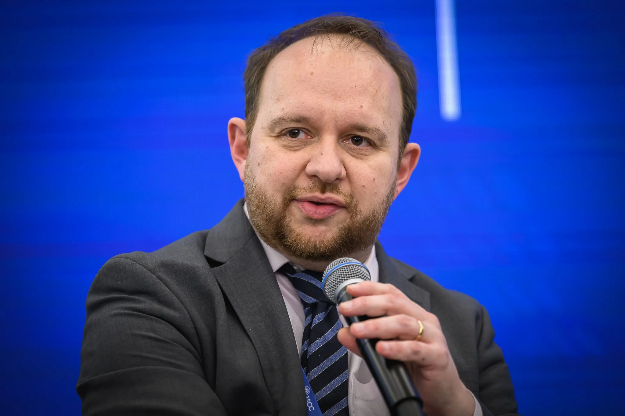 A man in a dark suit and striped tie holds a microphone during the panel discussion.