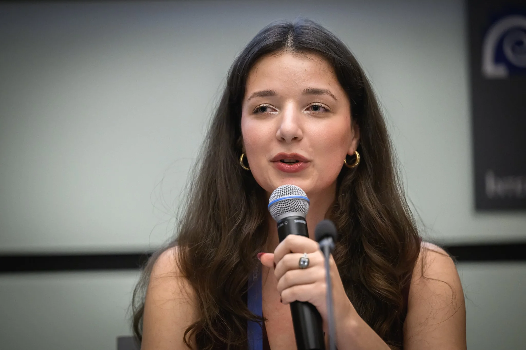 A woman with long dark hair speaks into a handheld microphone.