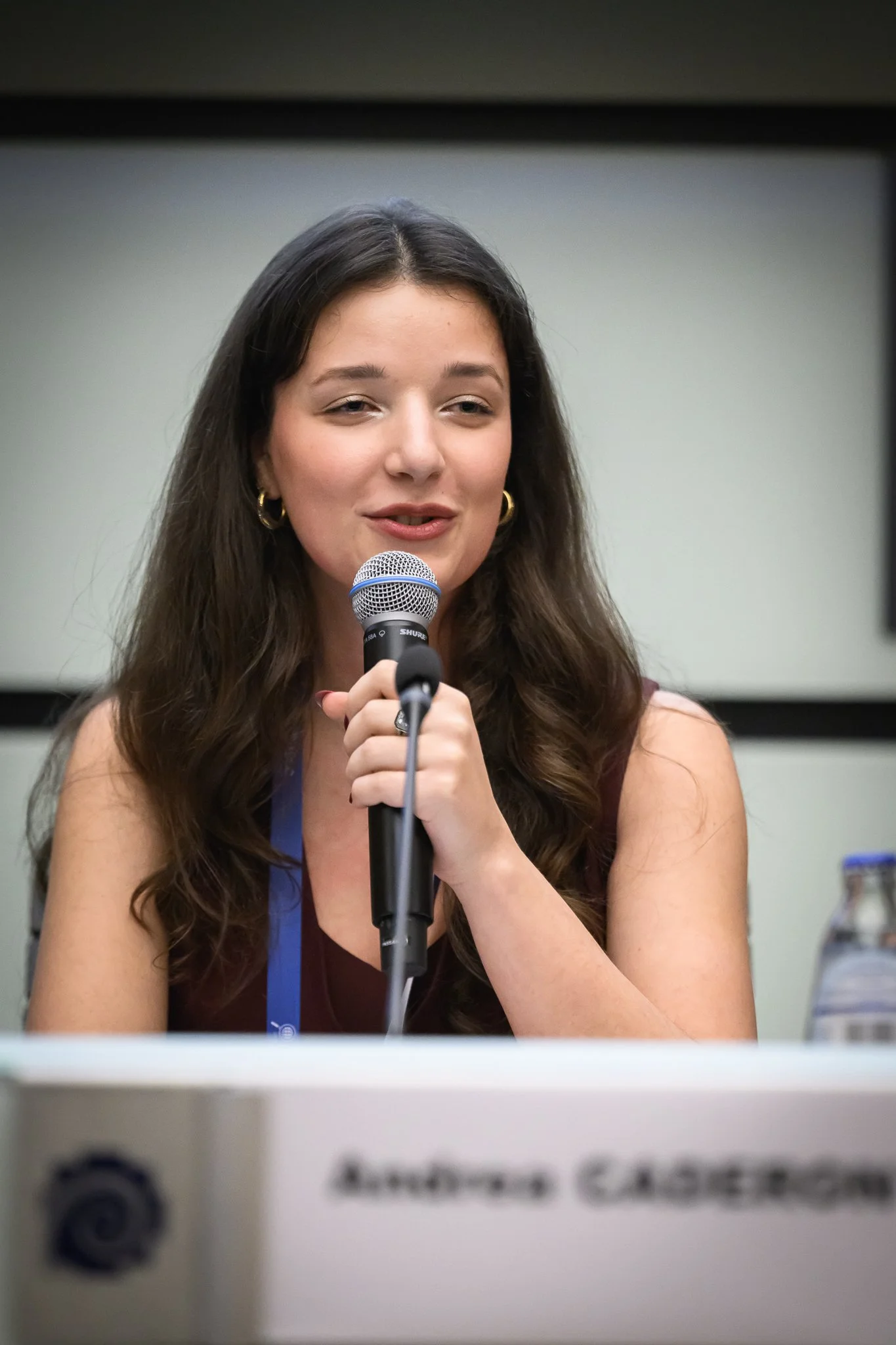 A woman speaks into a microphone behind a conference nameplate.