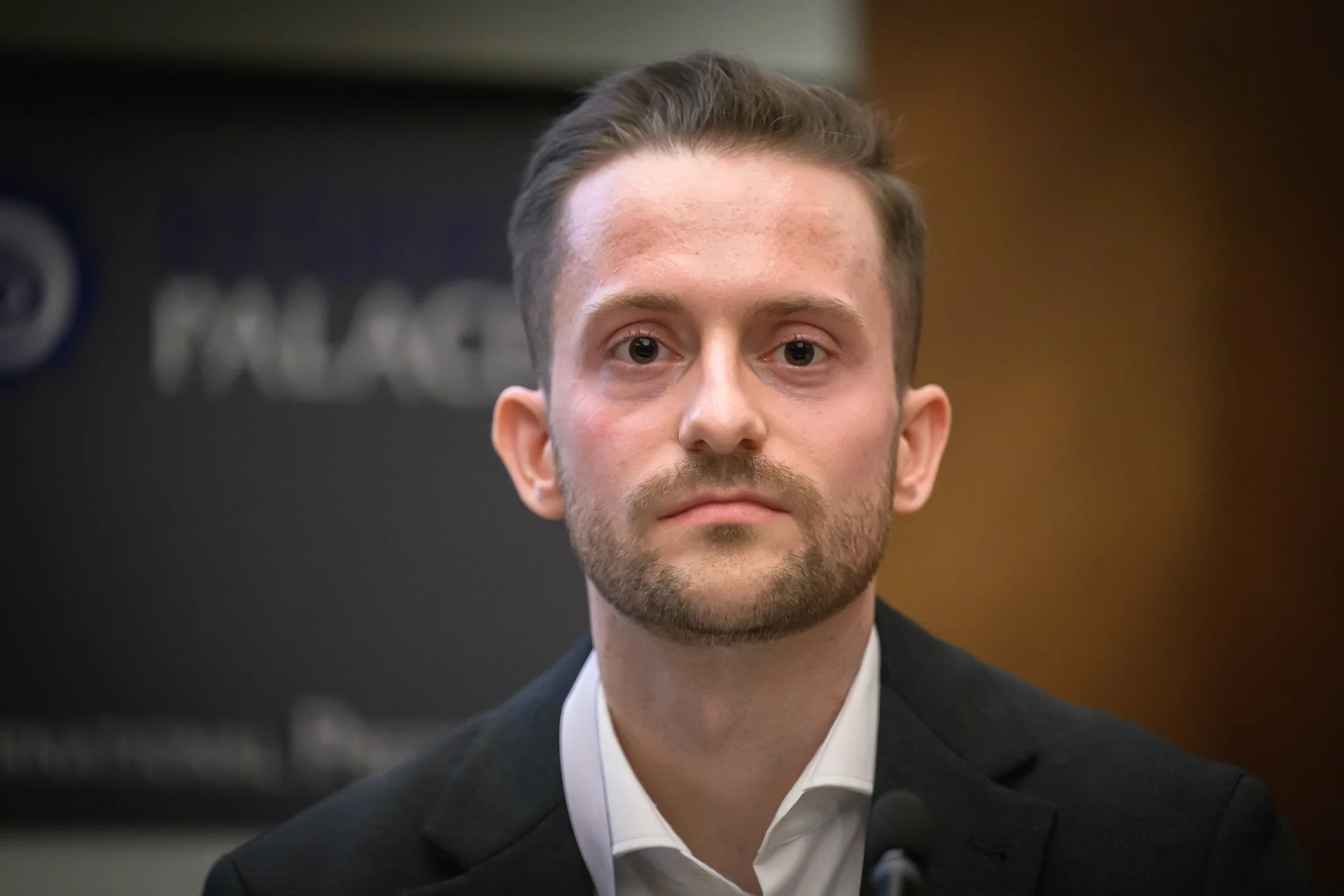 A man in a dark jacket and white shirt looks towards the speaker.