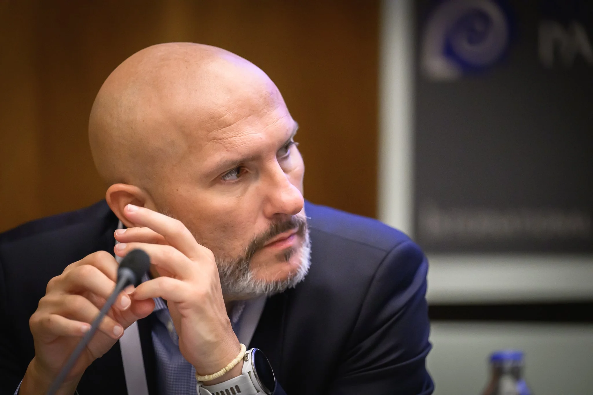 A bald man with a grey beard listens while holding his hands near a desk microphone.
