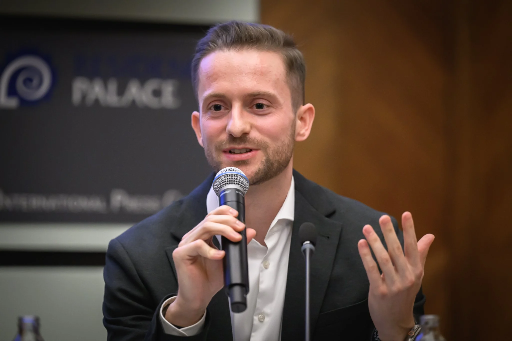 A man in a dark jacket gestures while speaking into a handheld microphone.