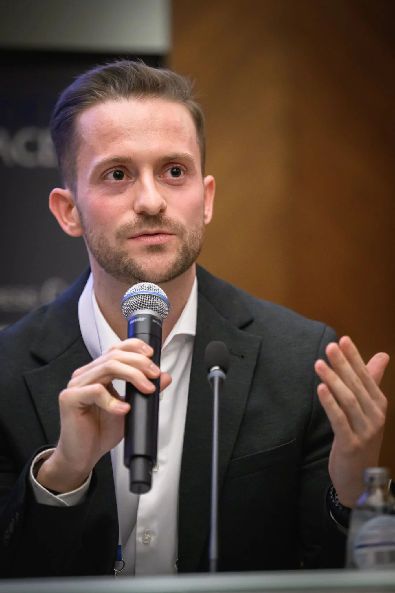 A man speaks into a microphone while gesturing with an open hand.