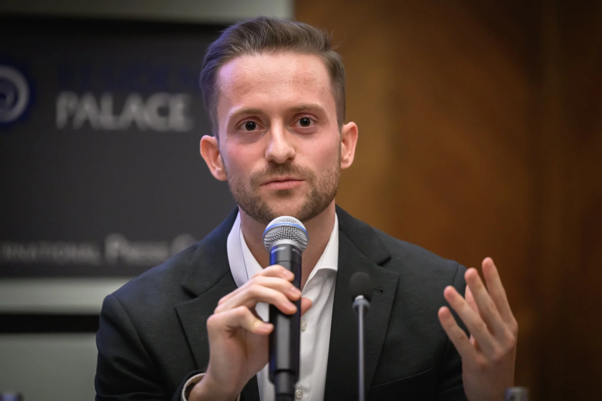 A seated man raises one hand while speaking into a microphone.