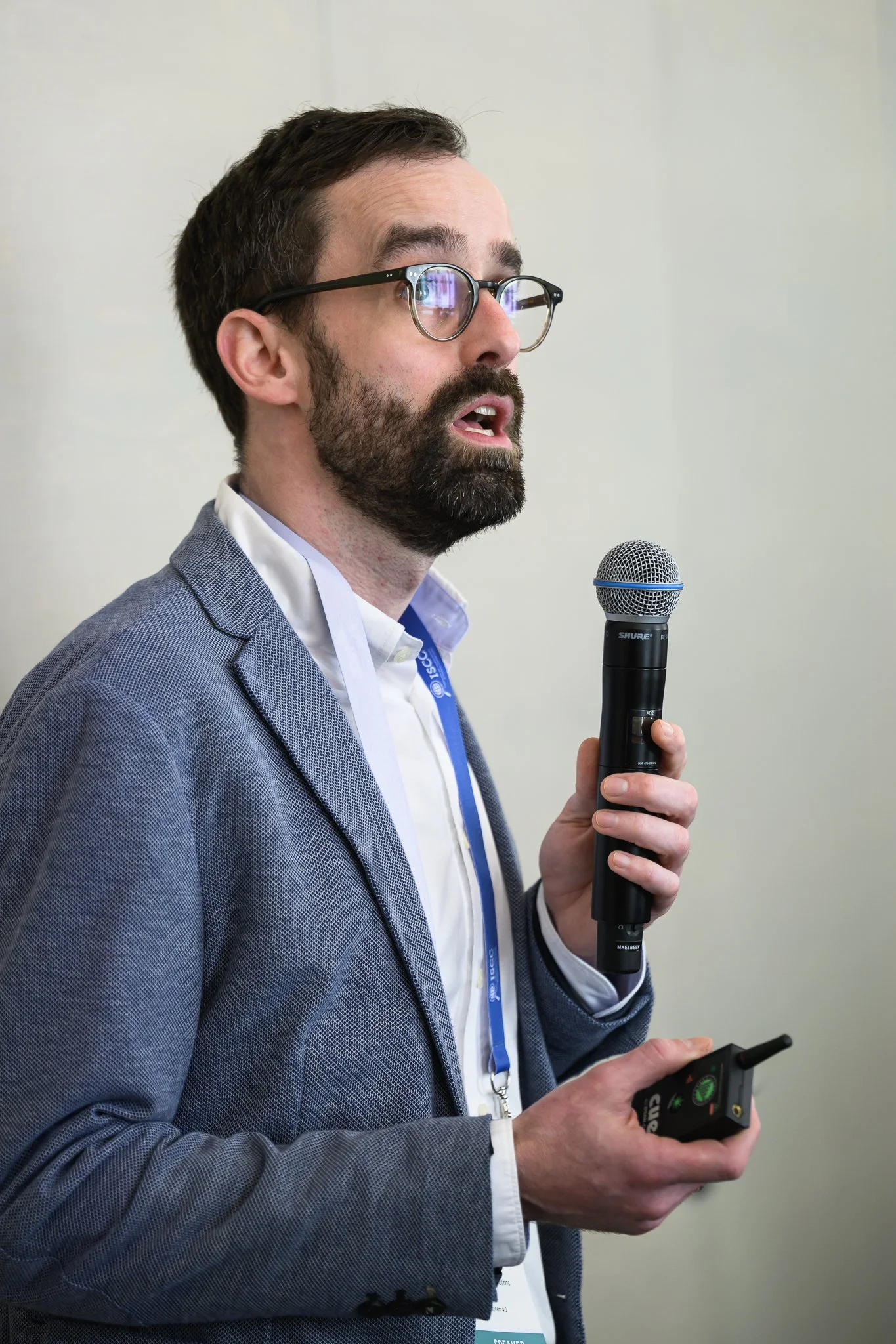A bearded presenter with glasses holds a microphone and remote control.