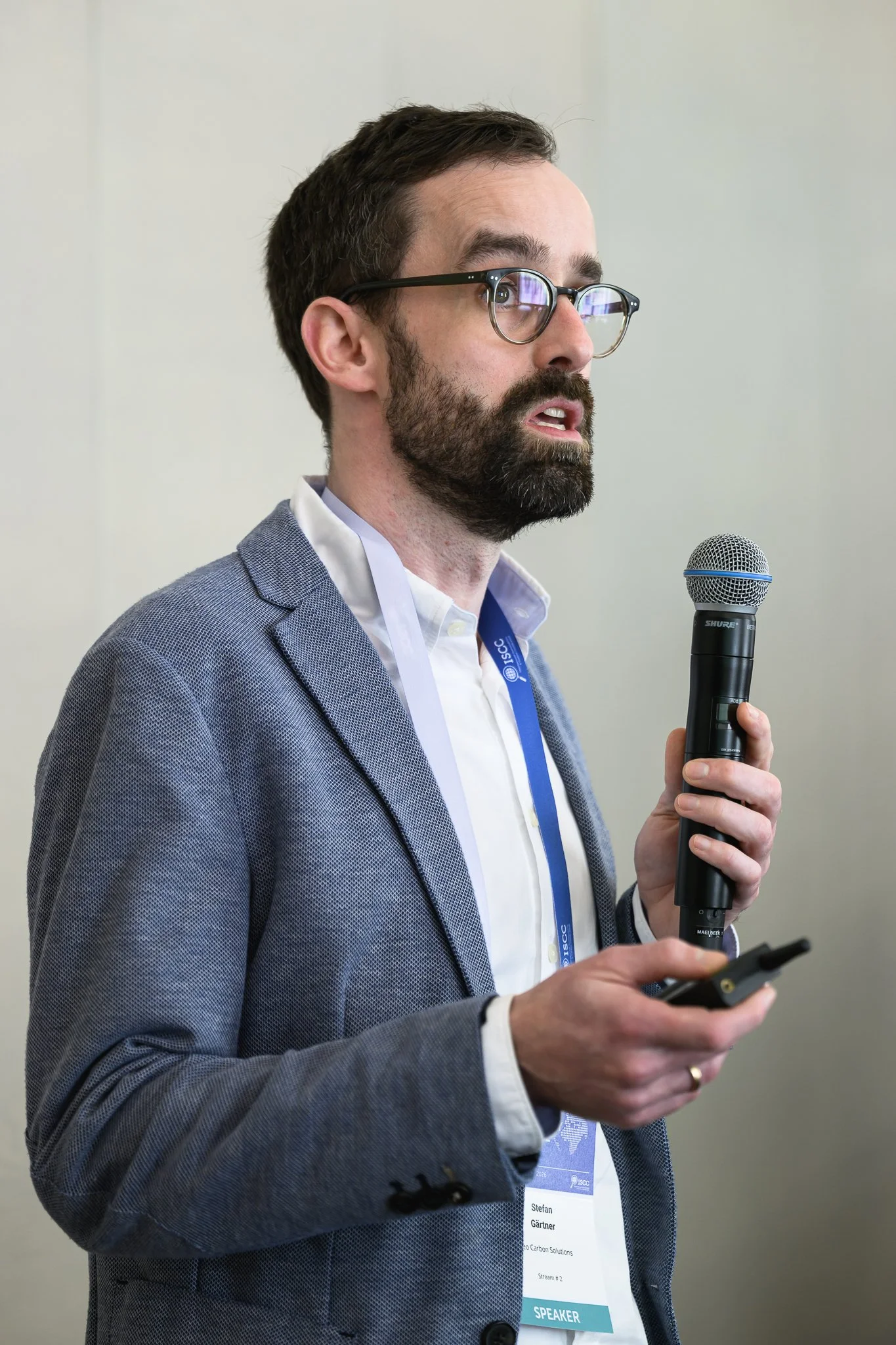 A man in a grey-blue jacket speaks into a handheld microphone.