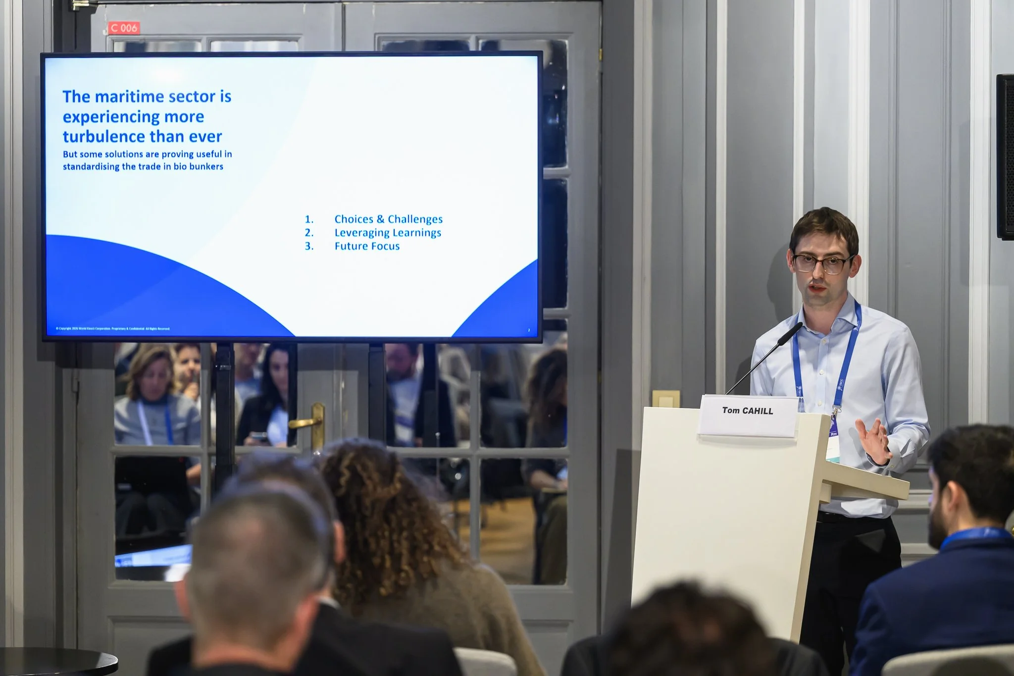 A man presents at a lectern beside a screen showing a slide about the maritime sector.
