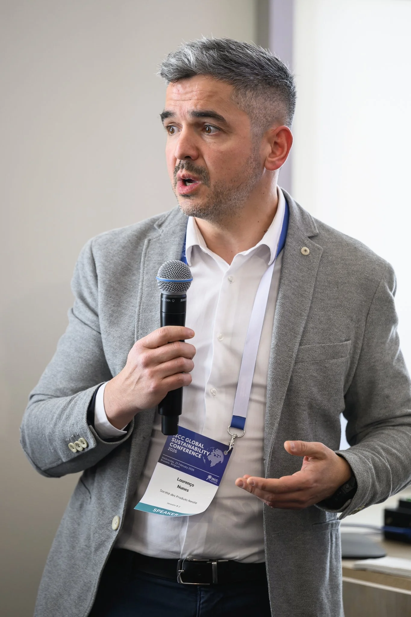 A man in a grey jacket speaks to the audience with one hand open.