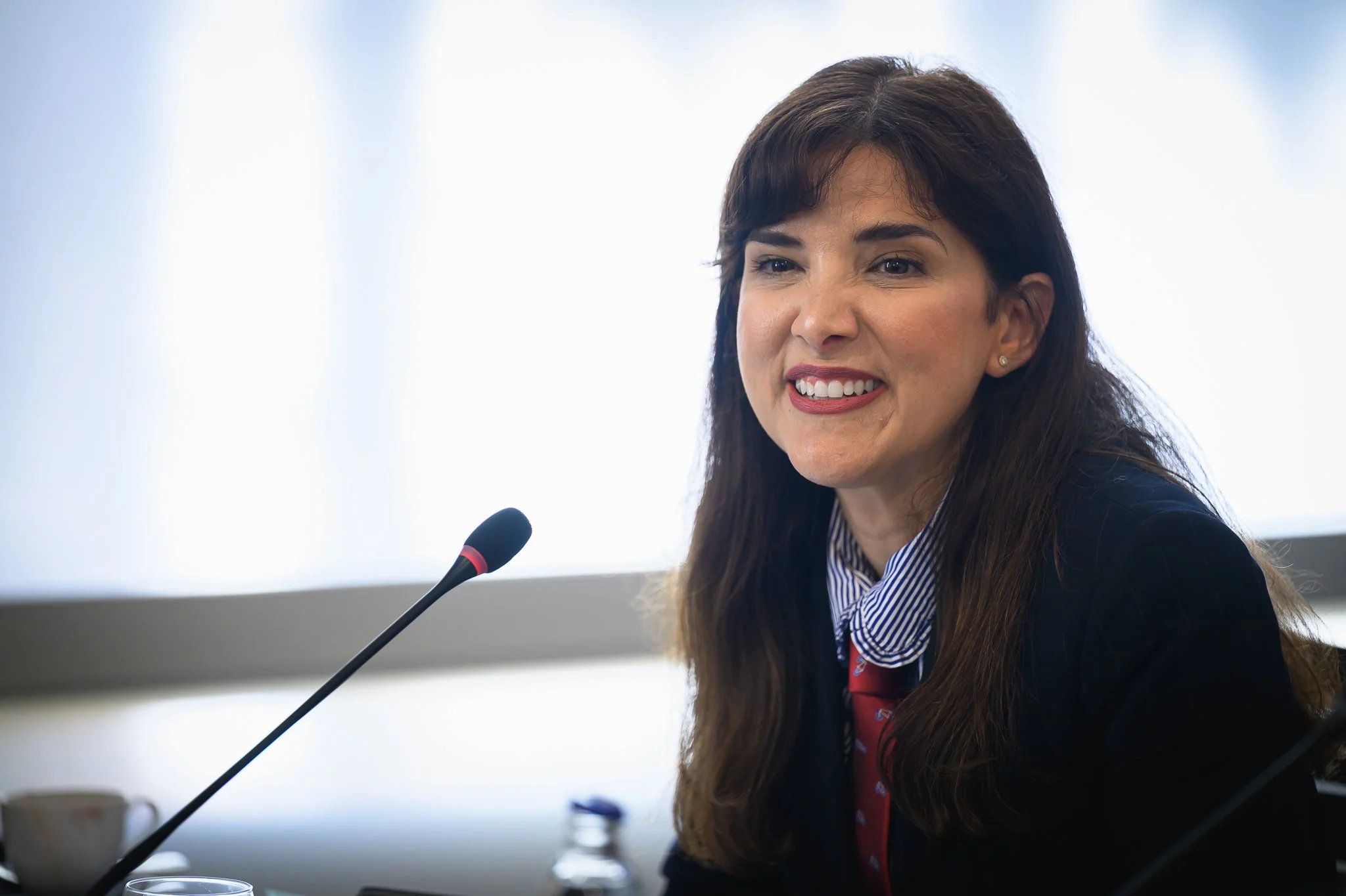 A smiling woman sits beside a desk microphone at a conference table.