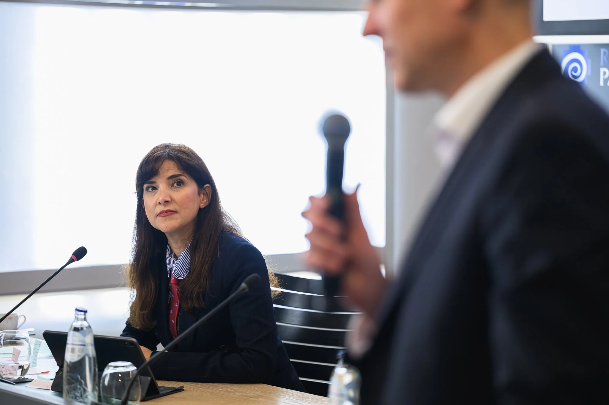 A seated woman listens as a man standing nearby speaks into a handheld microphone.