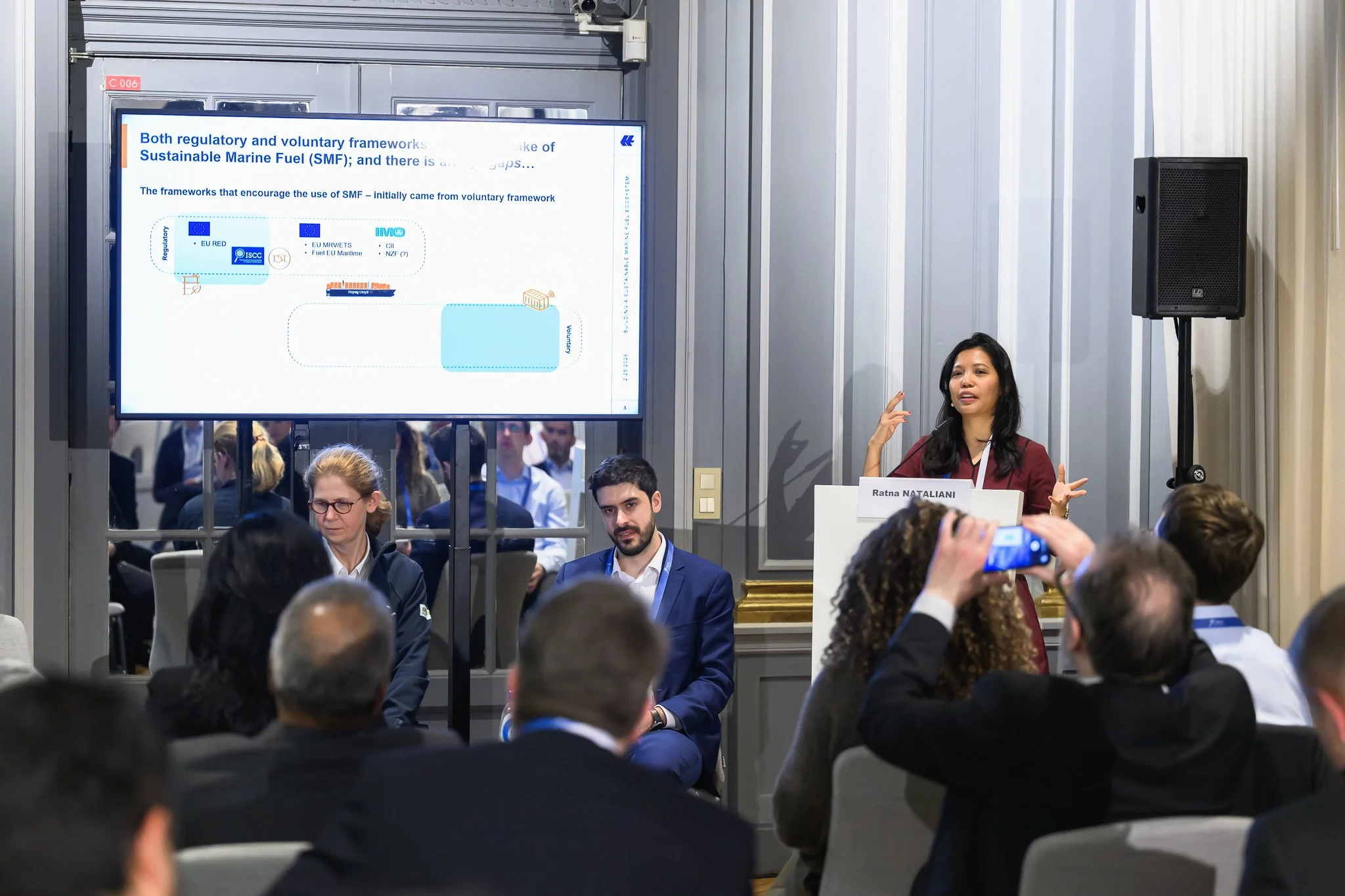 A seated audience watches a woman presenting beside a screen with a flow diagram.