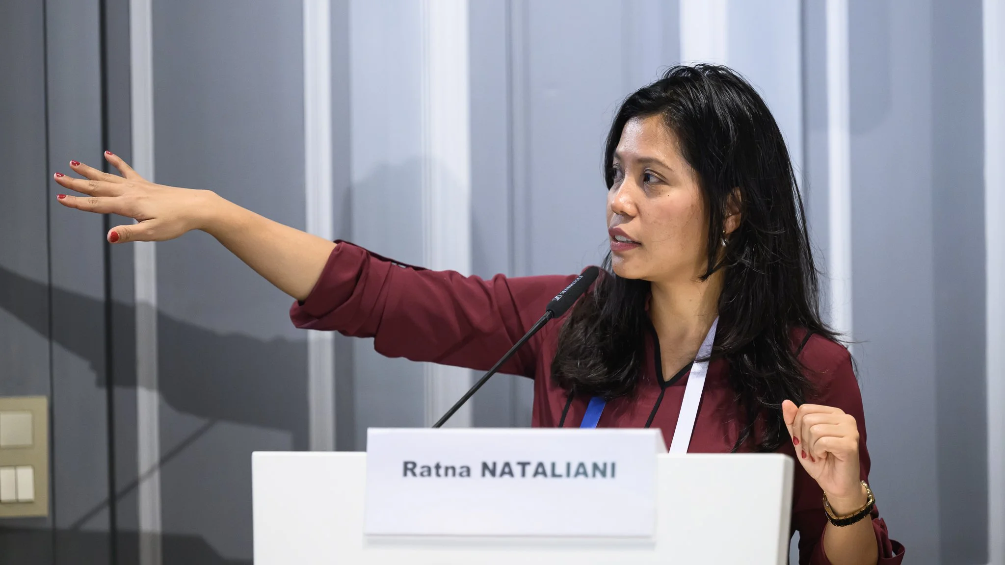Ratna Natalianti extends one arm towards the presentation screen while speaking.