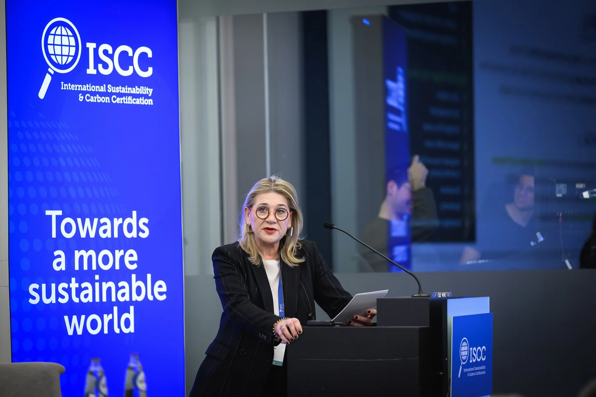 A woman with glasses stands at the lectern holding presentation notes.