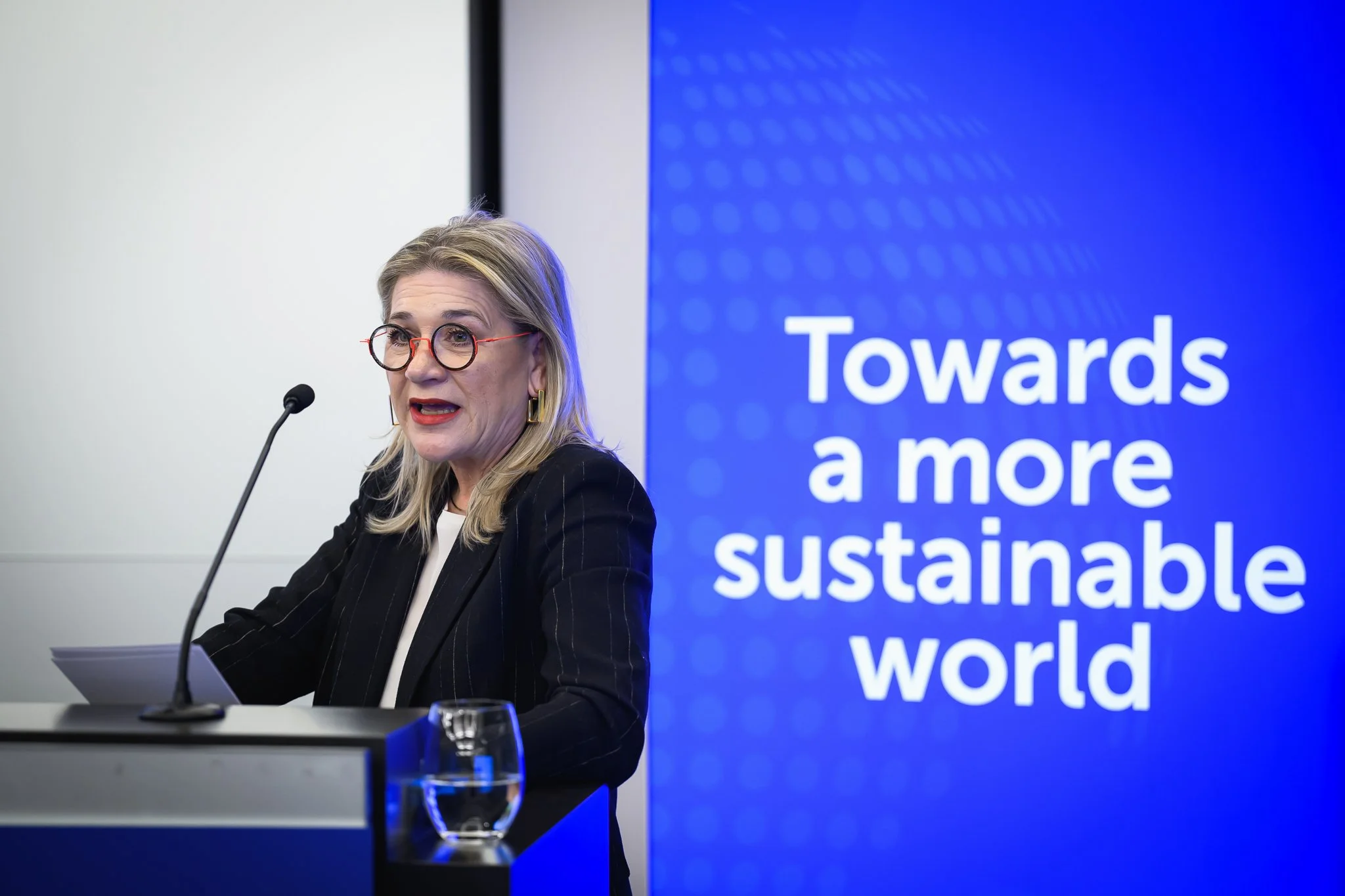 A woman with glasses speaks at the lectern beside an ISCC banner.