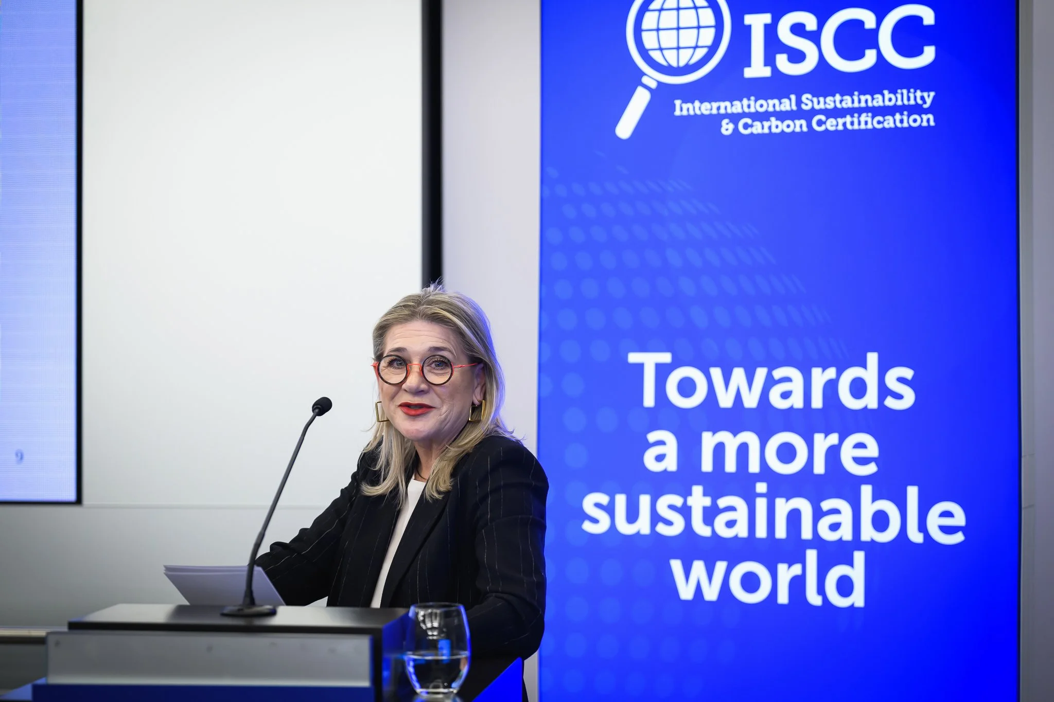 A woman with glasses addresses the conference beside a blue ISCC banner.