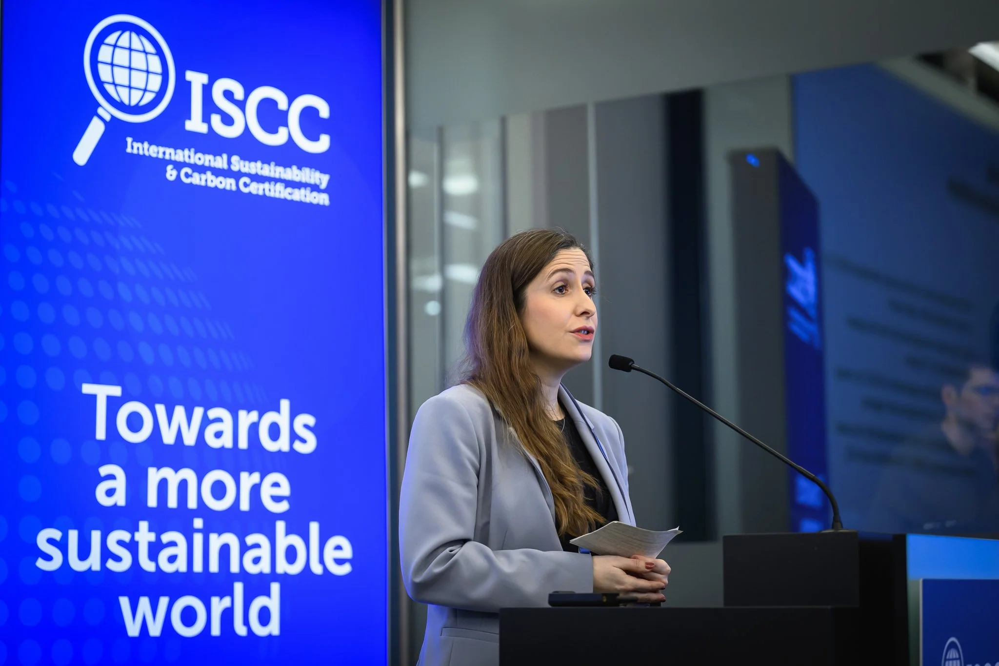 A woman in a light blue jacket holds notes while speaking at the lectern.