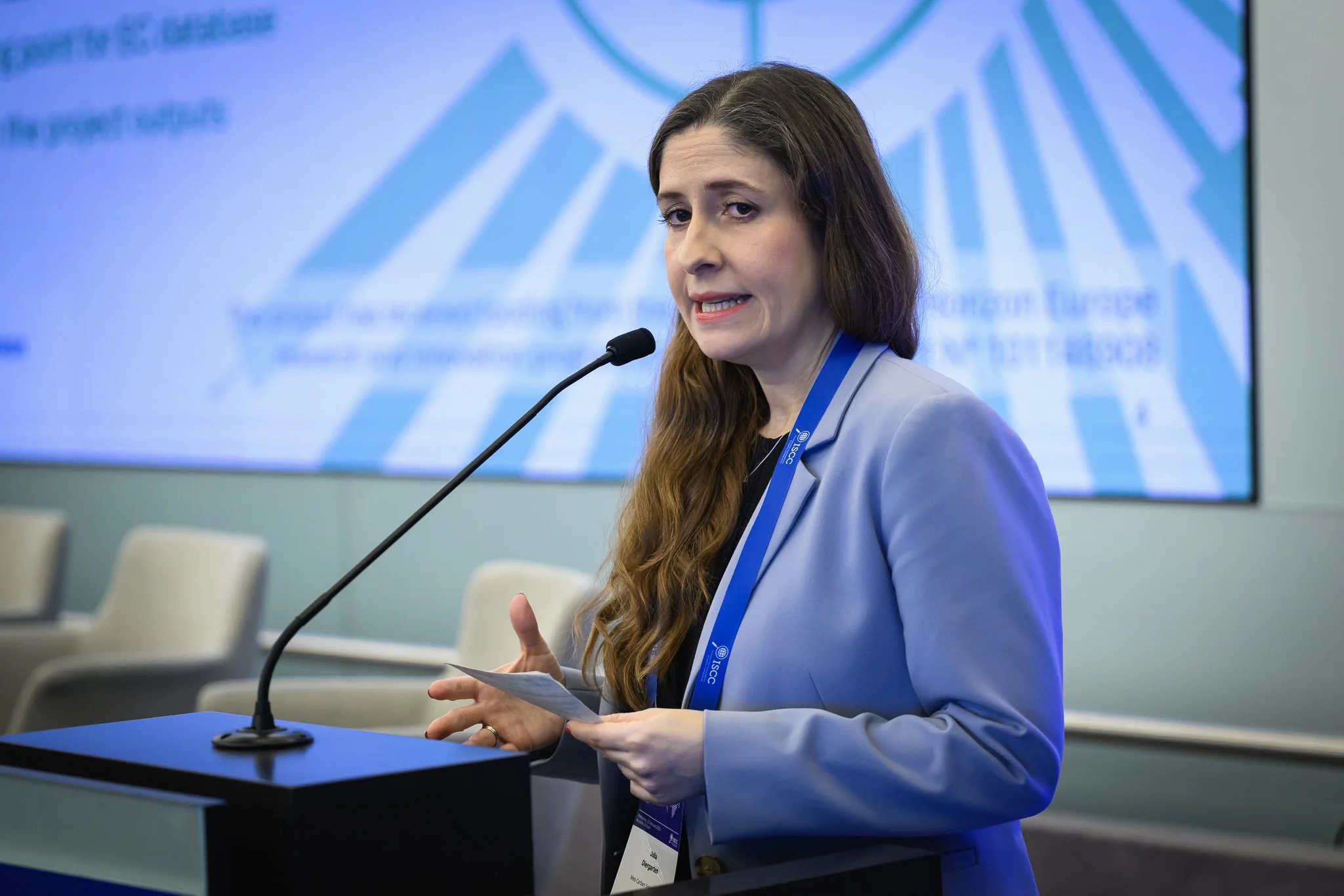 A woman in a light blue jacket presents in front of a projected slide.