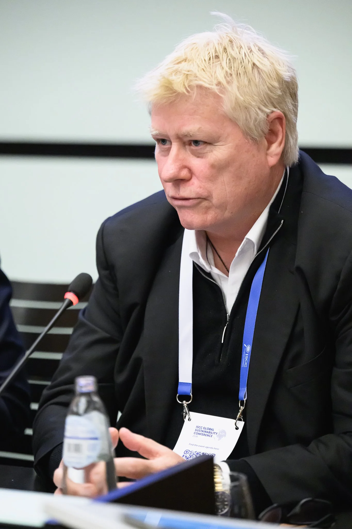 A man with light curly hair listens at a conference table beside a desk microphone.