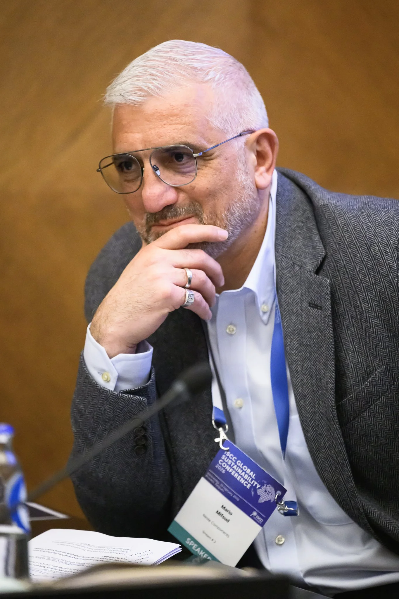 A bald man with glasses listens with one hand resting near his mouth.