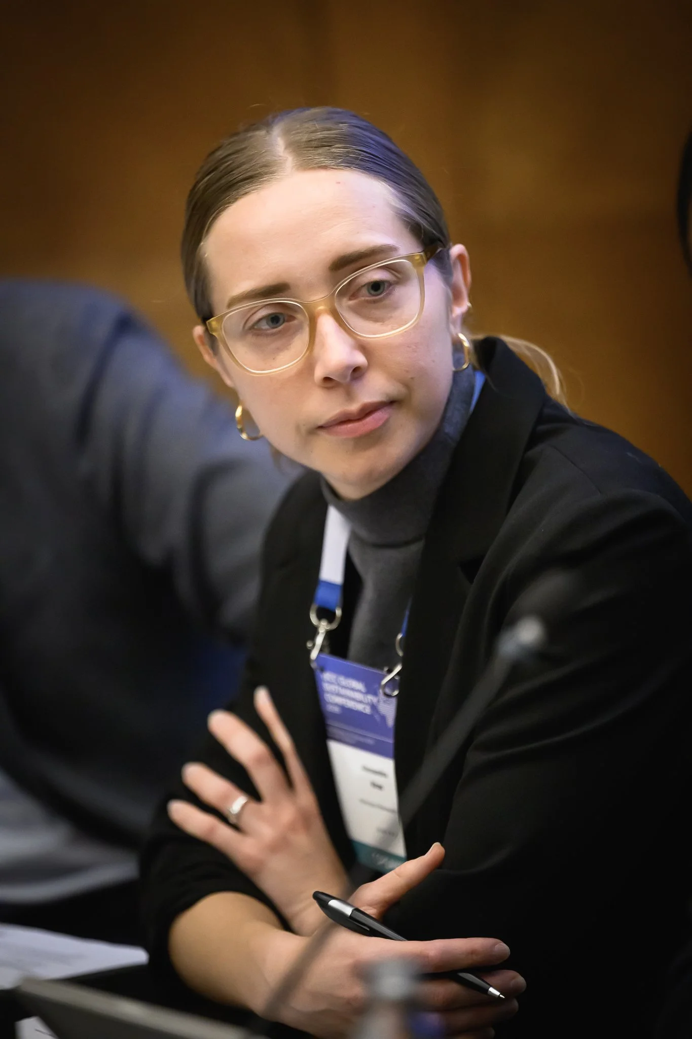 A woman with glasses listens with her arms folded during a conference session.