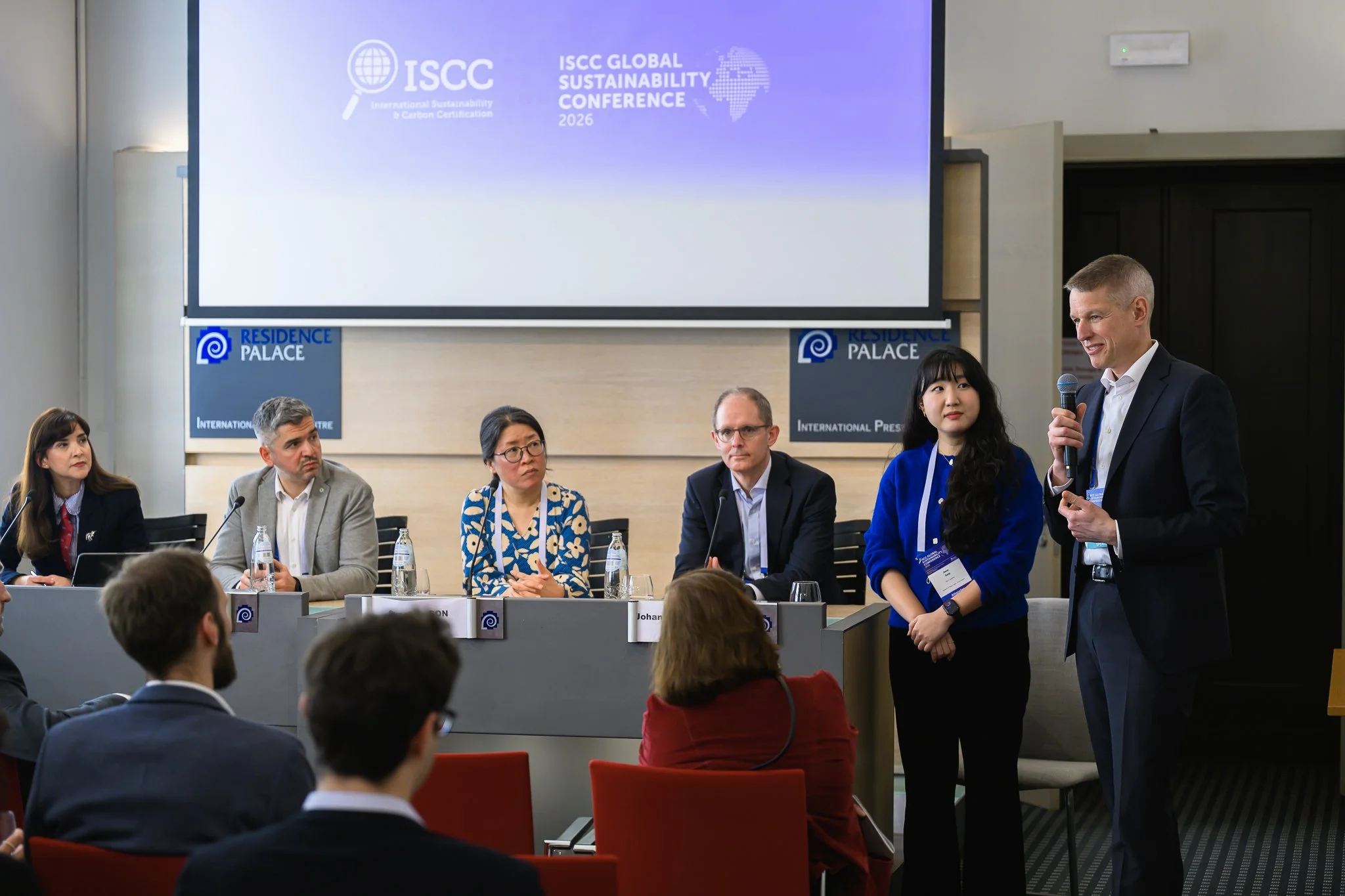 A moderator stands beside seated panel participants beneath a conference screen.