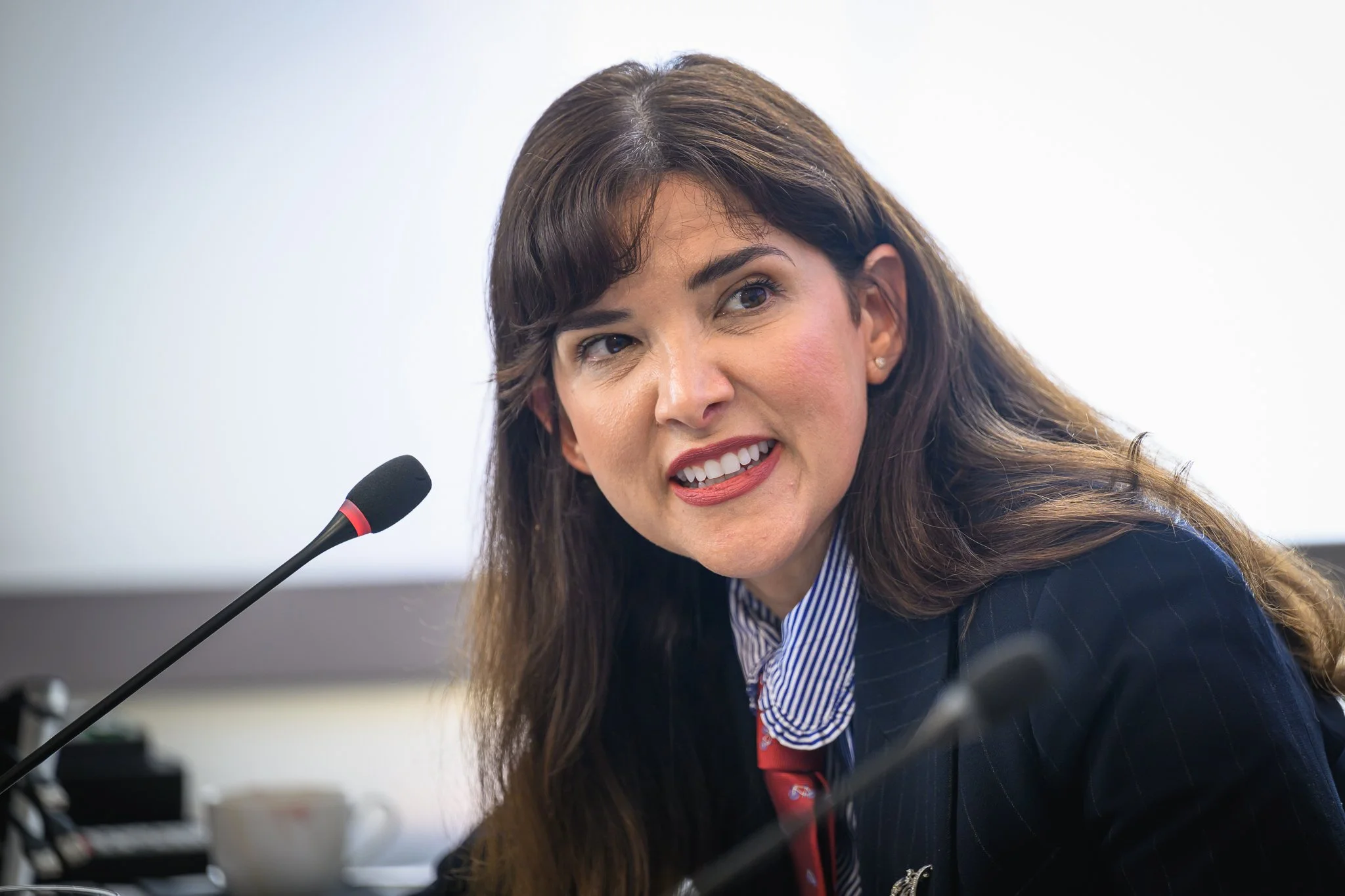 A smiling woman with long dark hair sits beside a desk microphone.