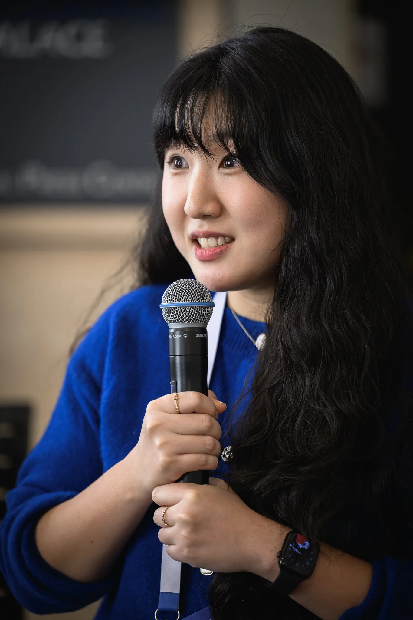 A smiling woman in a blue outfit holds a microphone during a panel discussion.