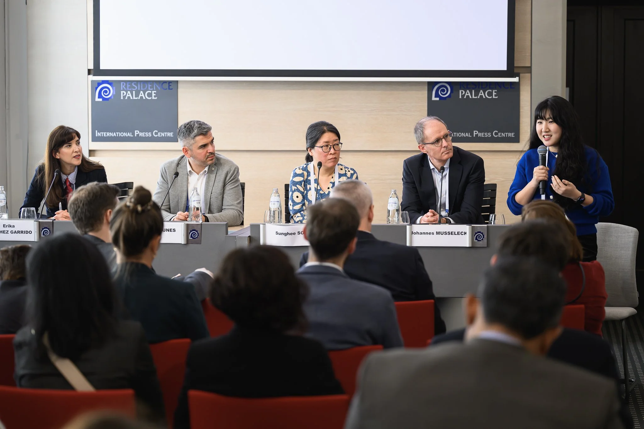 Five participants sit at a panel table while the audience listens.