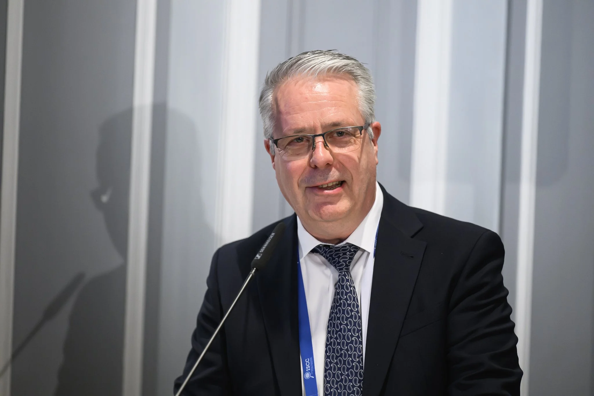 A man with grey hair and glasses smiles while speaking at a desk microphone.