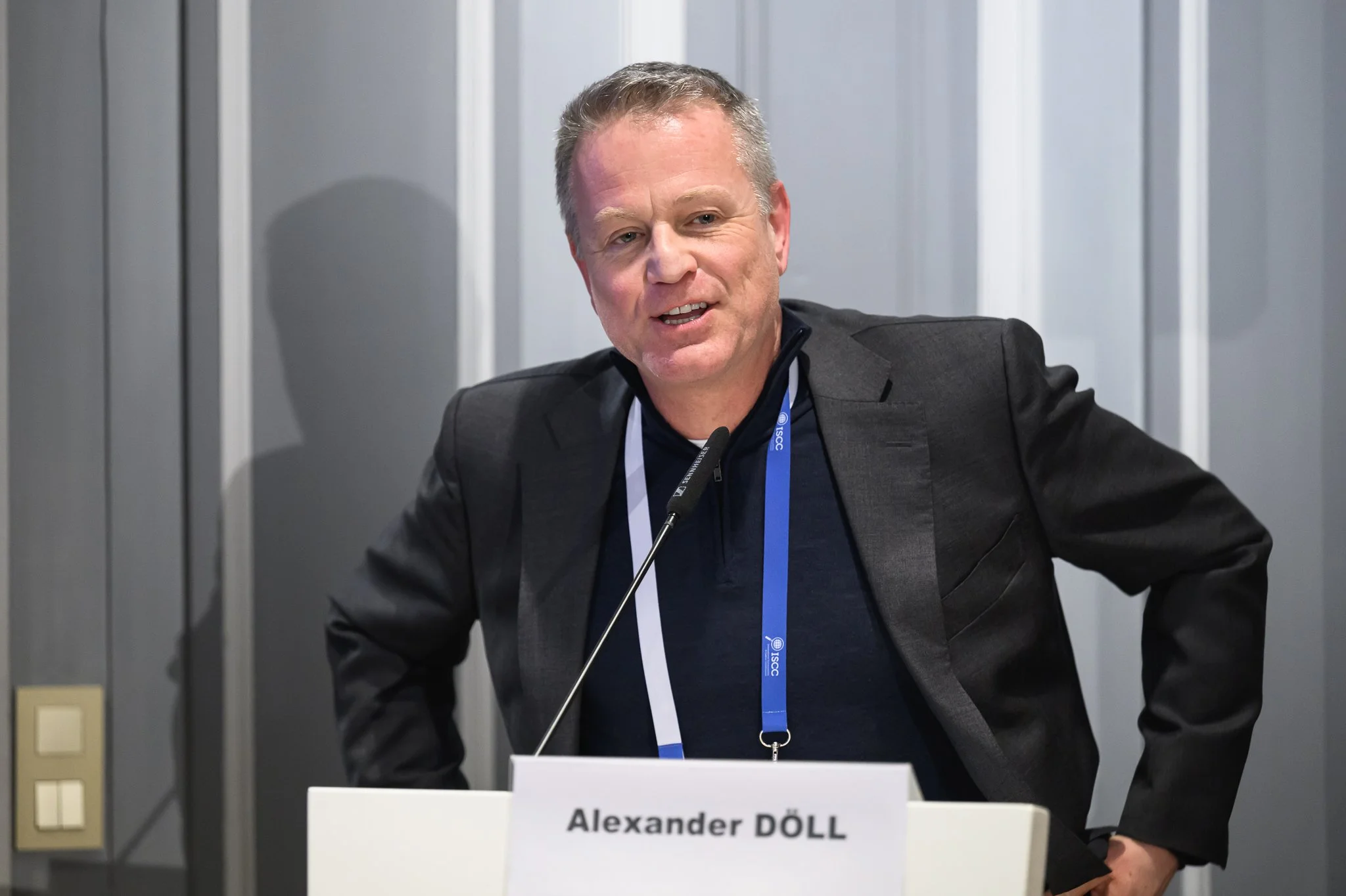 Alexander Doll speaks at the lectern with his hands resting near his hips.