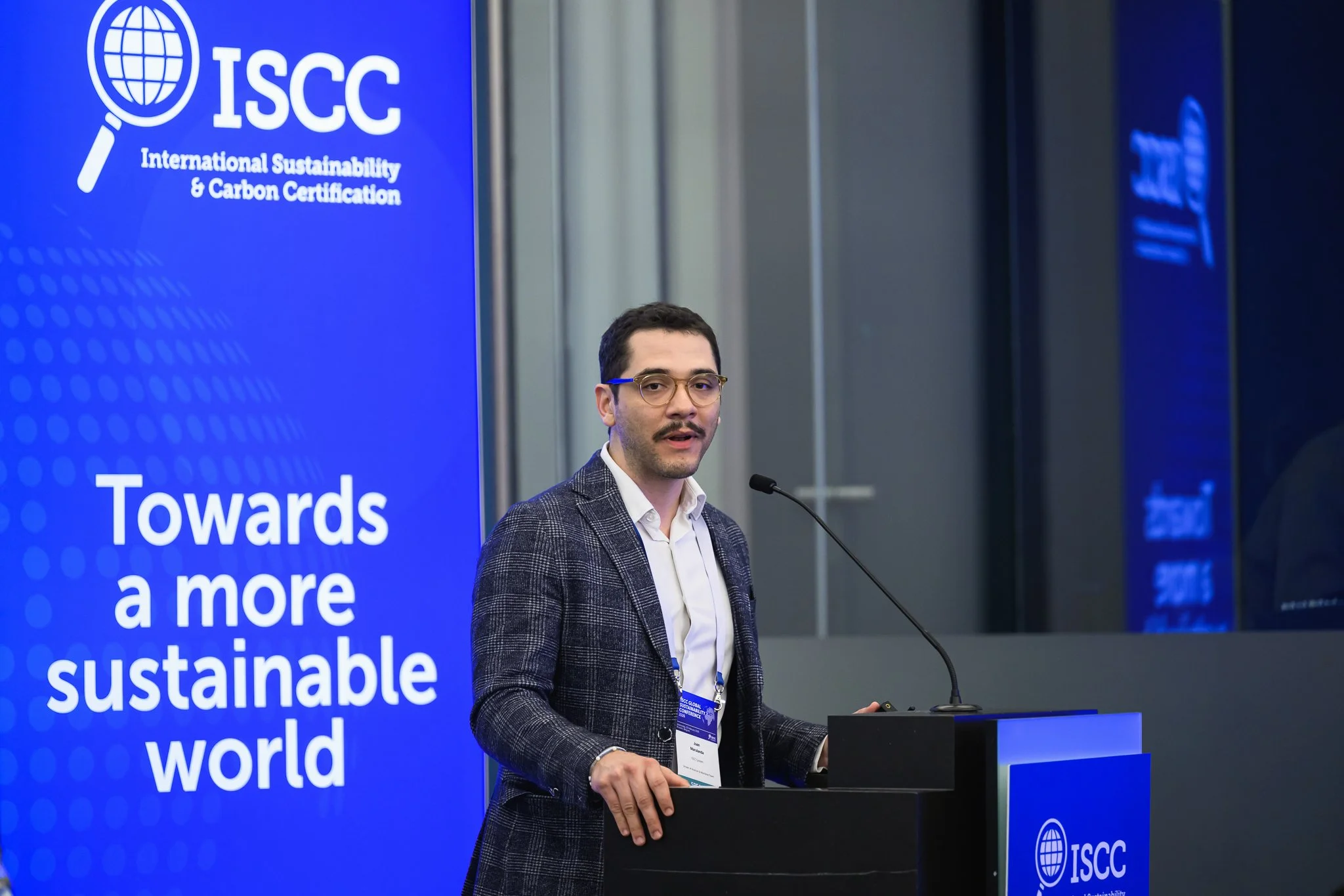 A man with glasses and a checked jacket stands at the lectern beside an ISCC banner.
