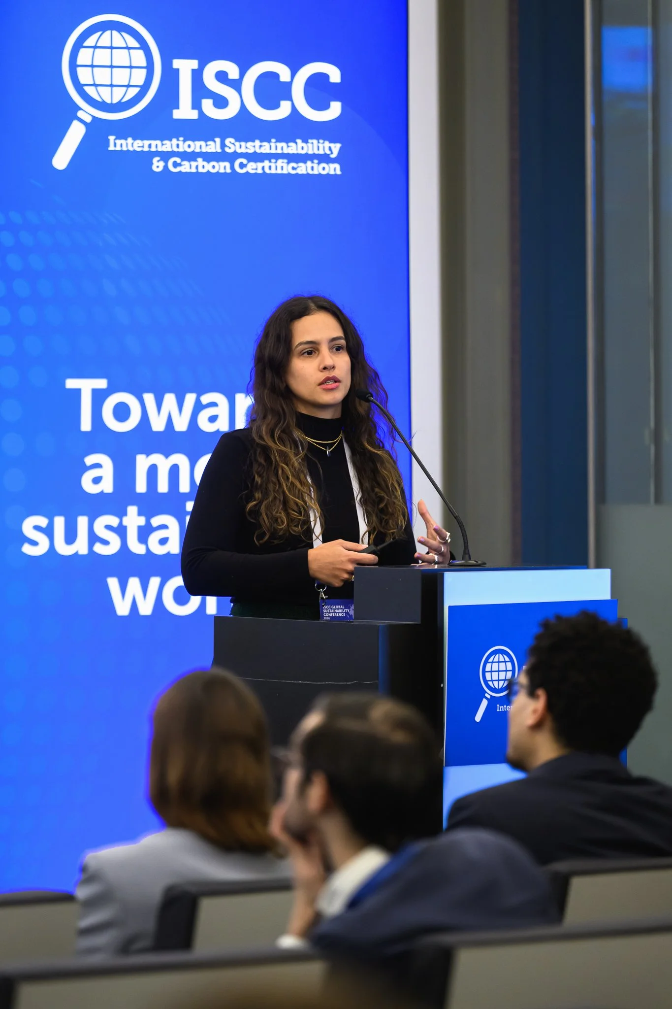 A woman addresses seated participants beside a blue ISCC banner.