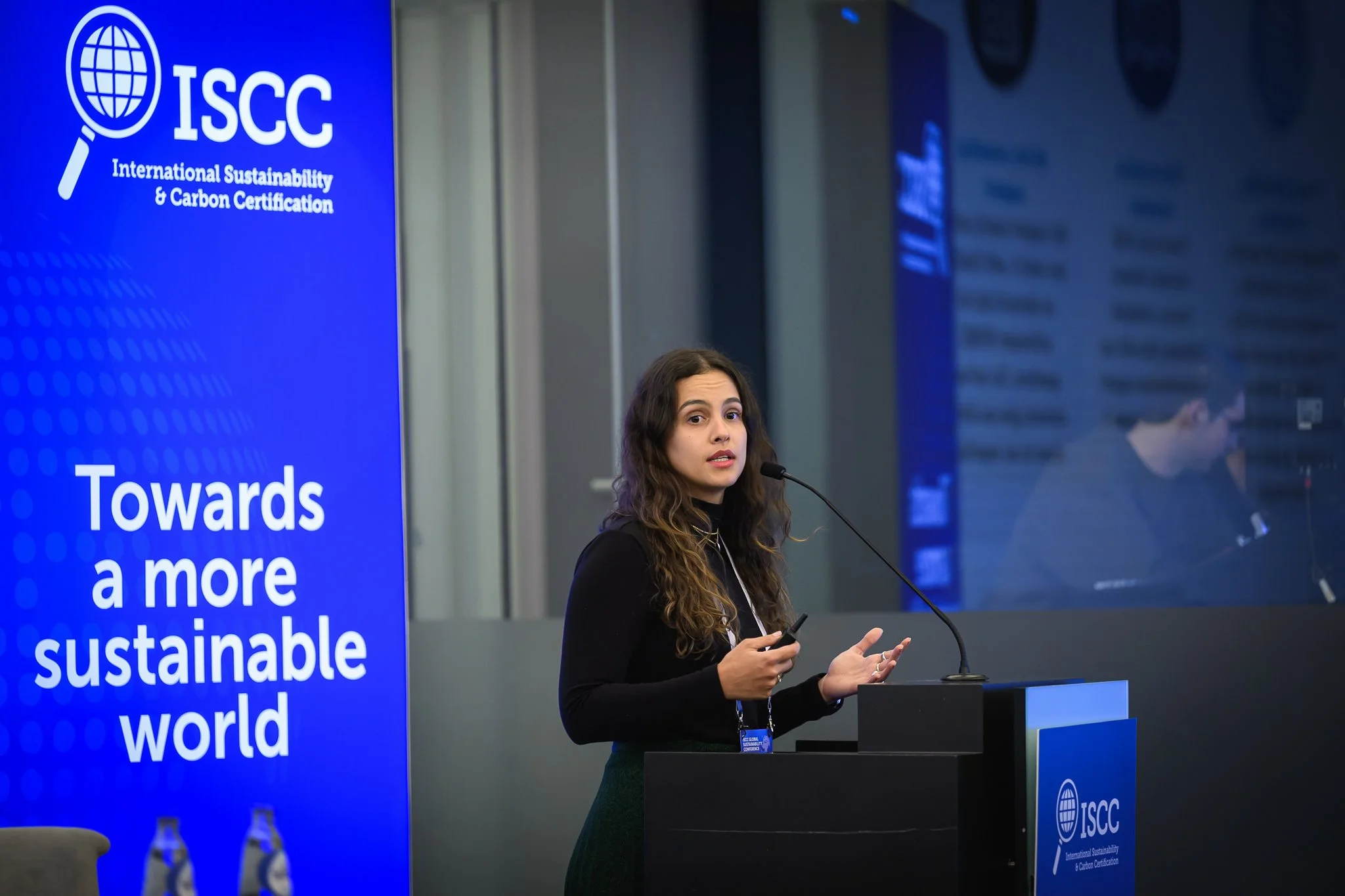 A woman with long curly hair gestures while speaking at the lectern.