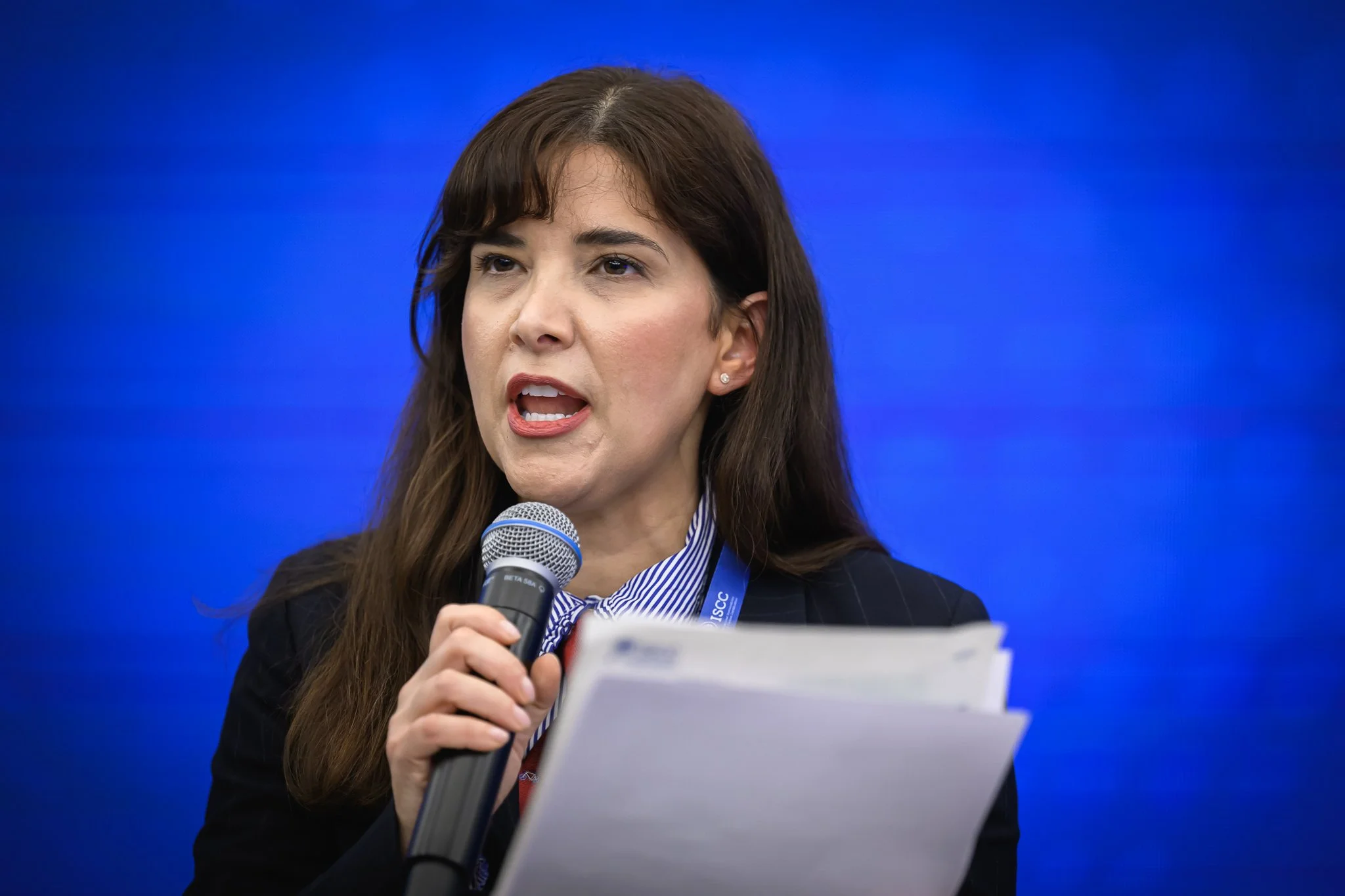 A woman with dark hair speaks into a microphone while holding printed notes.