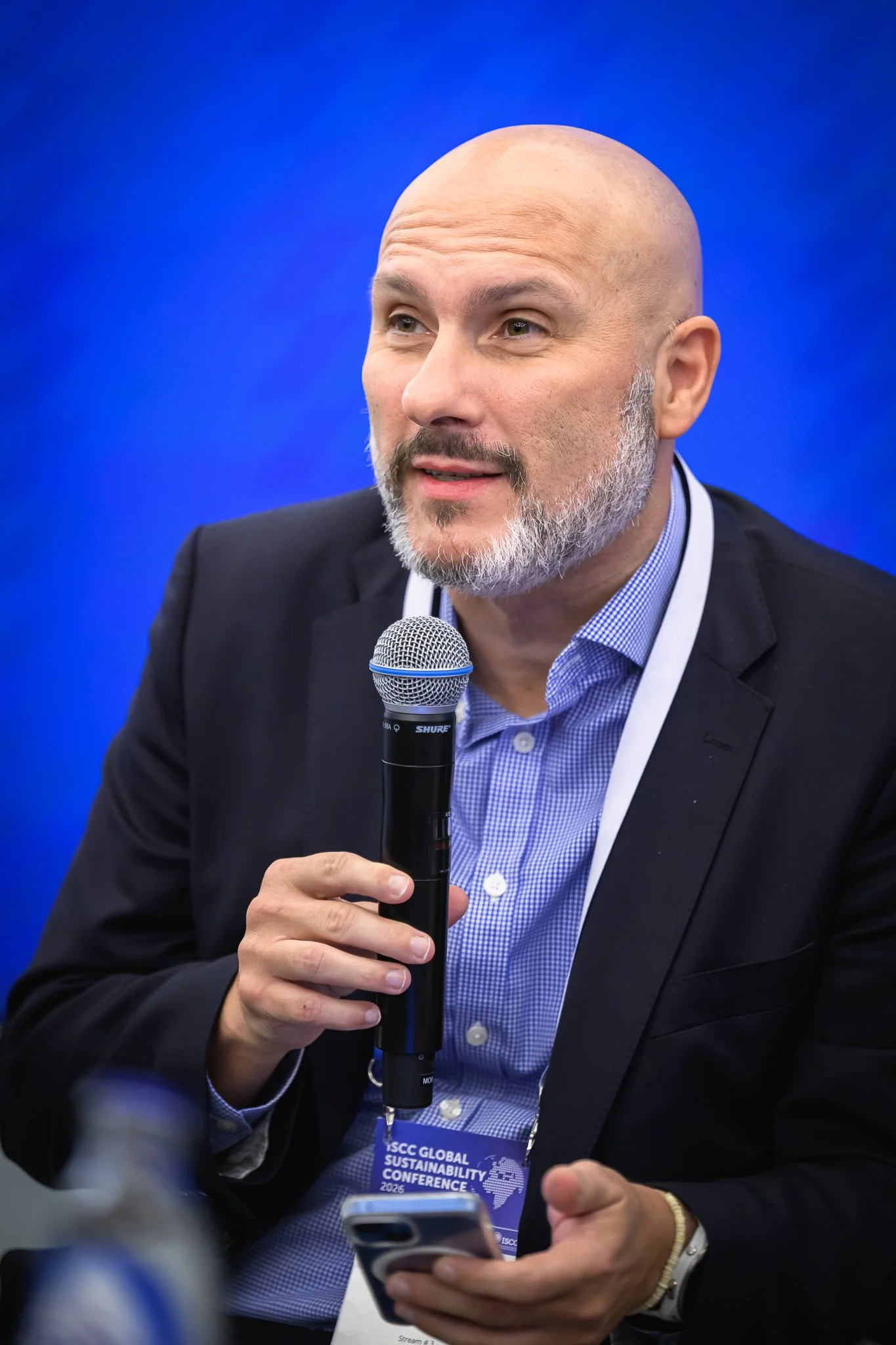 A bald panel participant speaks into a microphone while holding a smartphone.