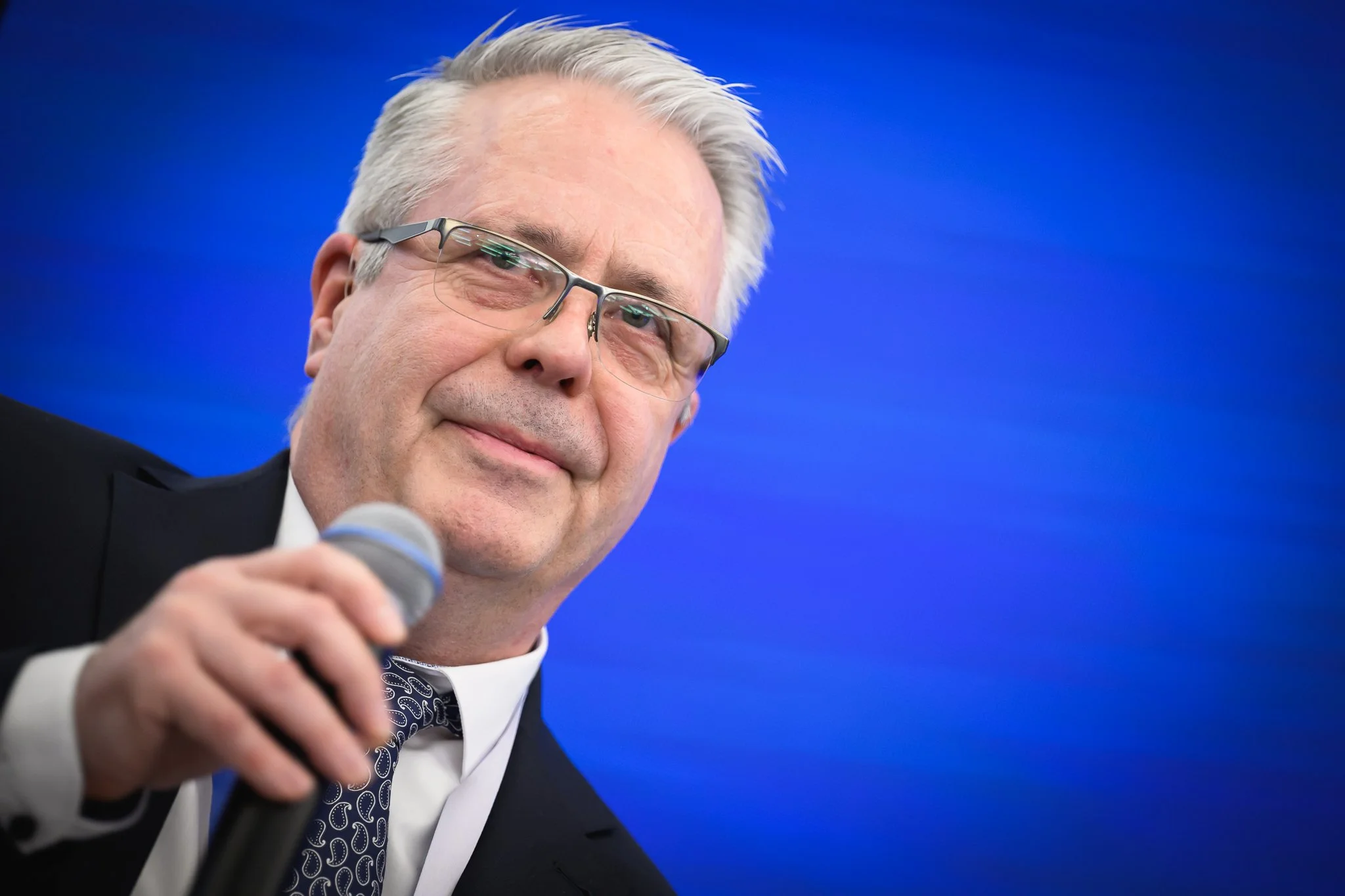 Close-up of an older man with glasses holding a microphone against a blue background.