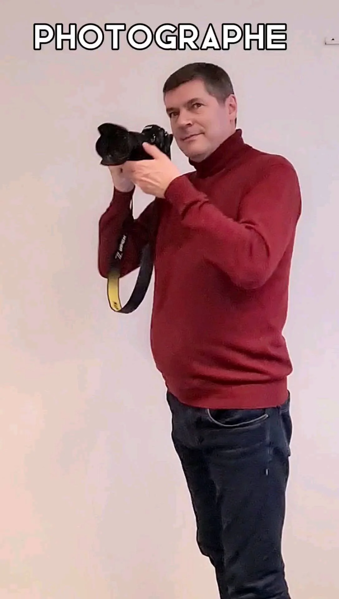 Un homme en pull rouge tient un appareil photo devant un mur clair, avec le mot PHOTOGRAPHE en surimpression.